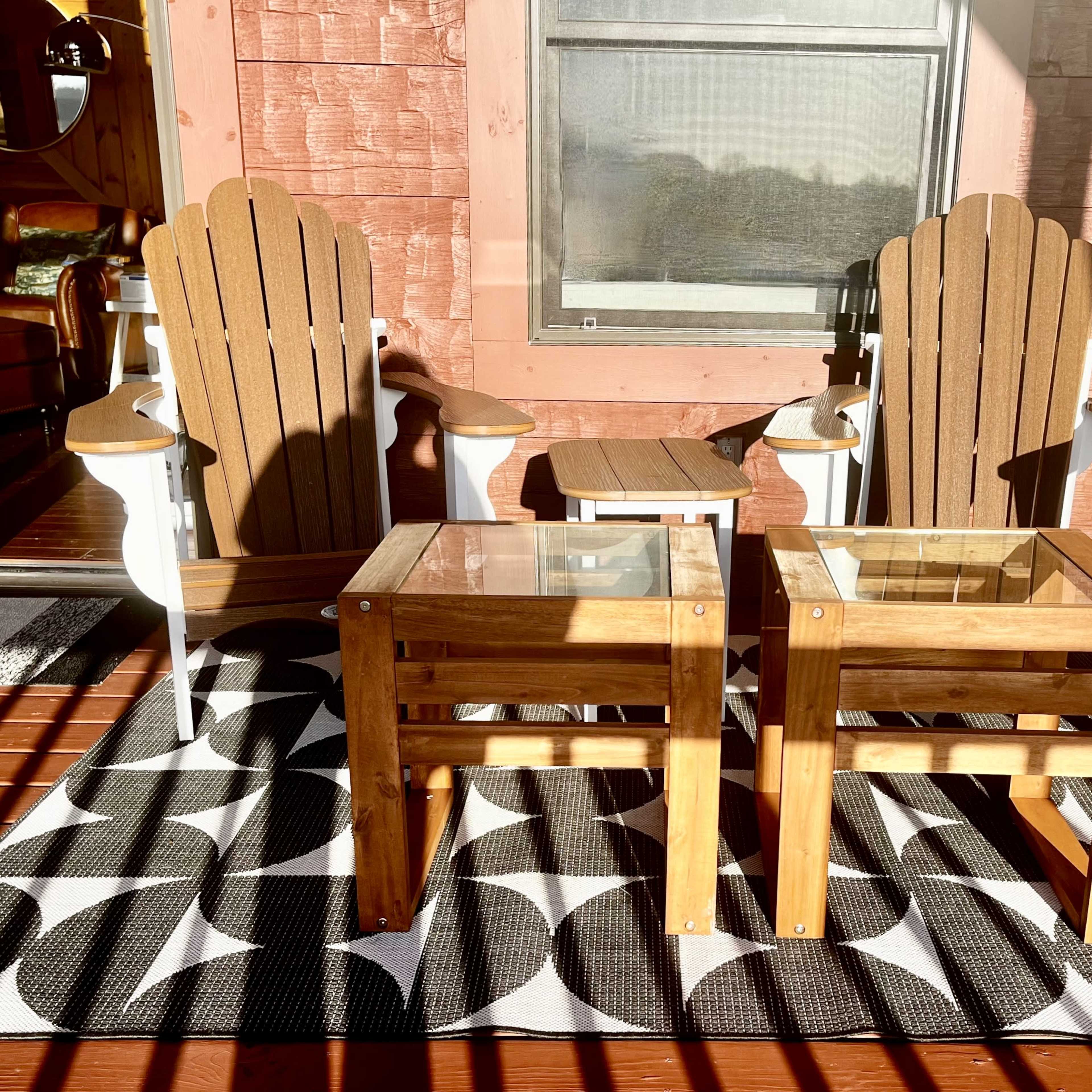 Two wooden Adirondack chairs are placed beside two square wooden tables on a patterned rug outdoors.