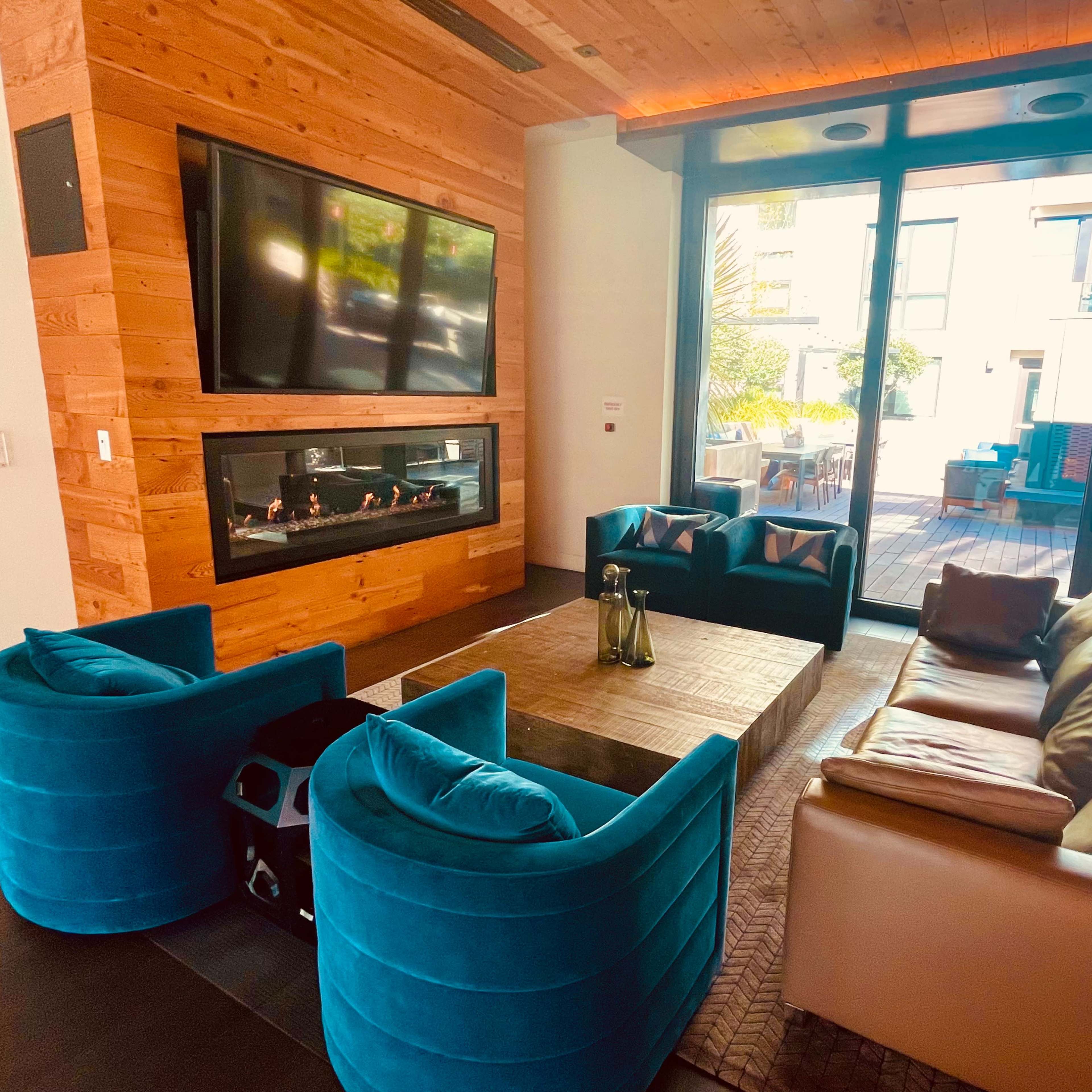 A modern living space with a wooden wall, a large window facing an outdoor area, and a seating arrangement that includes blue chairs and a brown sofa surrounding a wooden coffee table.