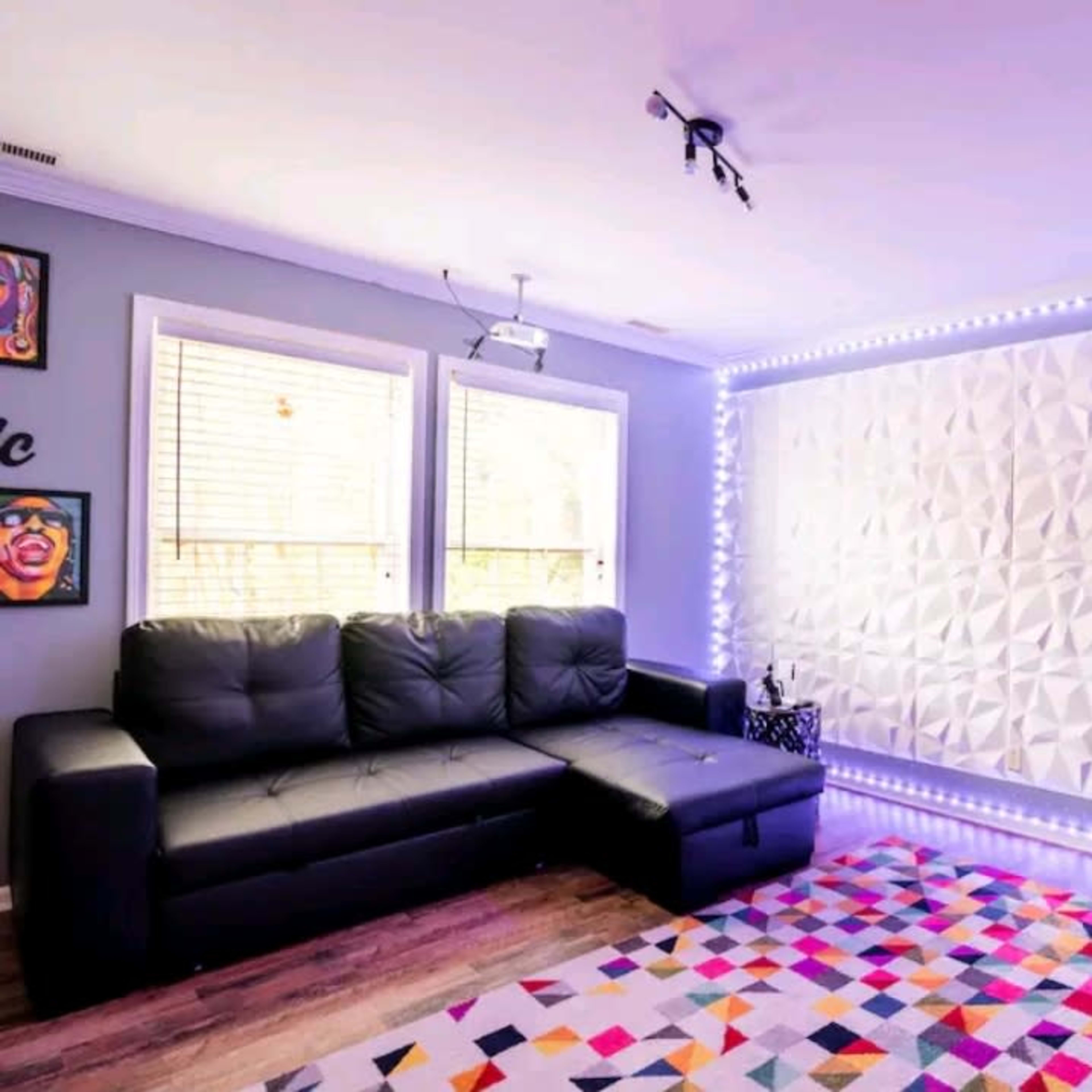 A modern living room features a sectional black sofa, a black armchair, a patterned area rug, and a textured wall illuminated with LED lights.