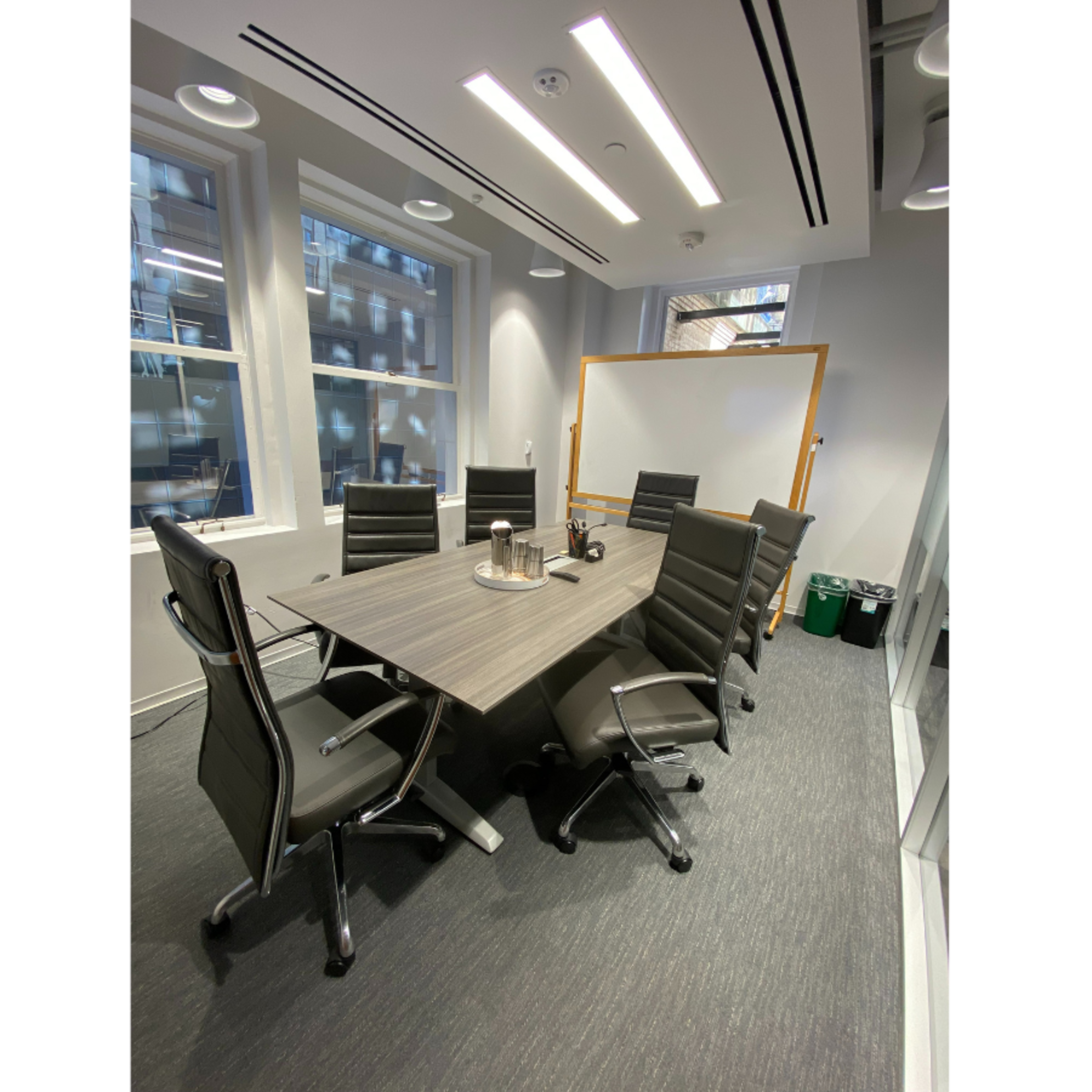 A modern conference room features a long table surrounded by ergonomic chairs, with large windows and a whiteboard in the background.