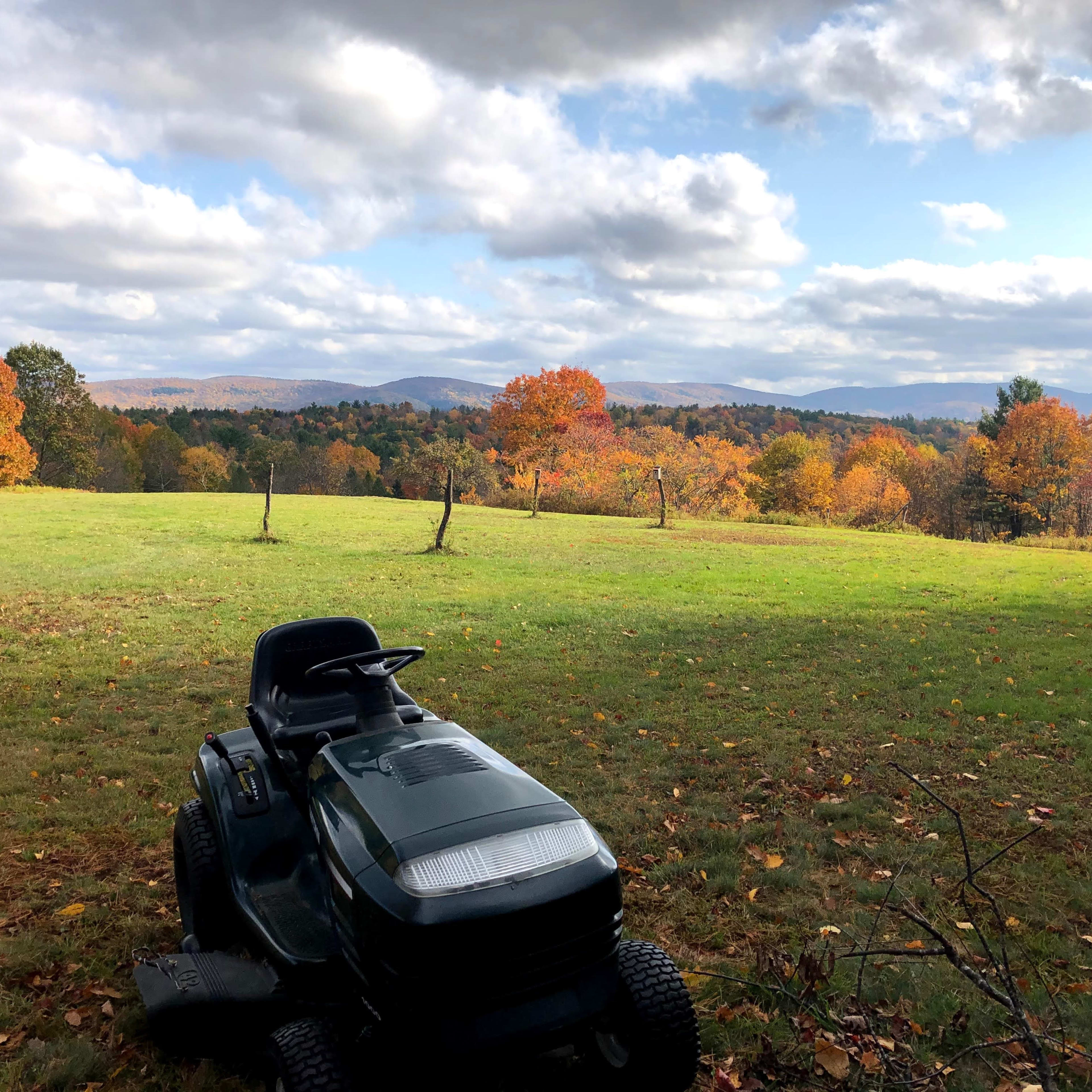 A lawn mower sits in a grassy area overlooking a landscape of autumn trees and distant mountains under a cloudy sky.