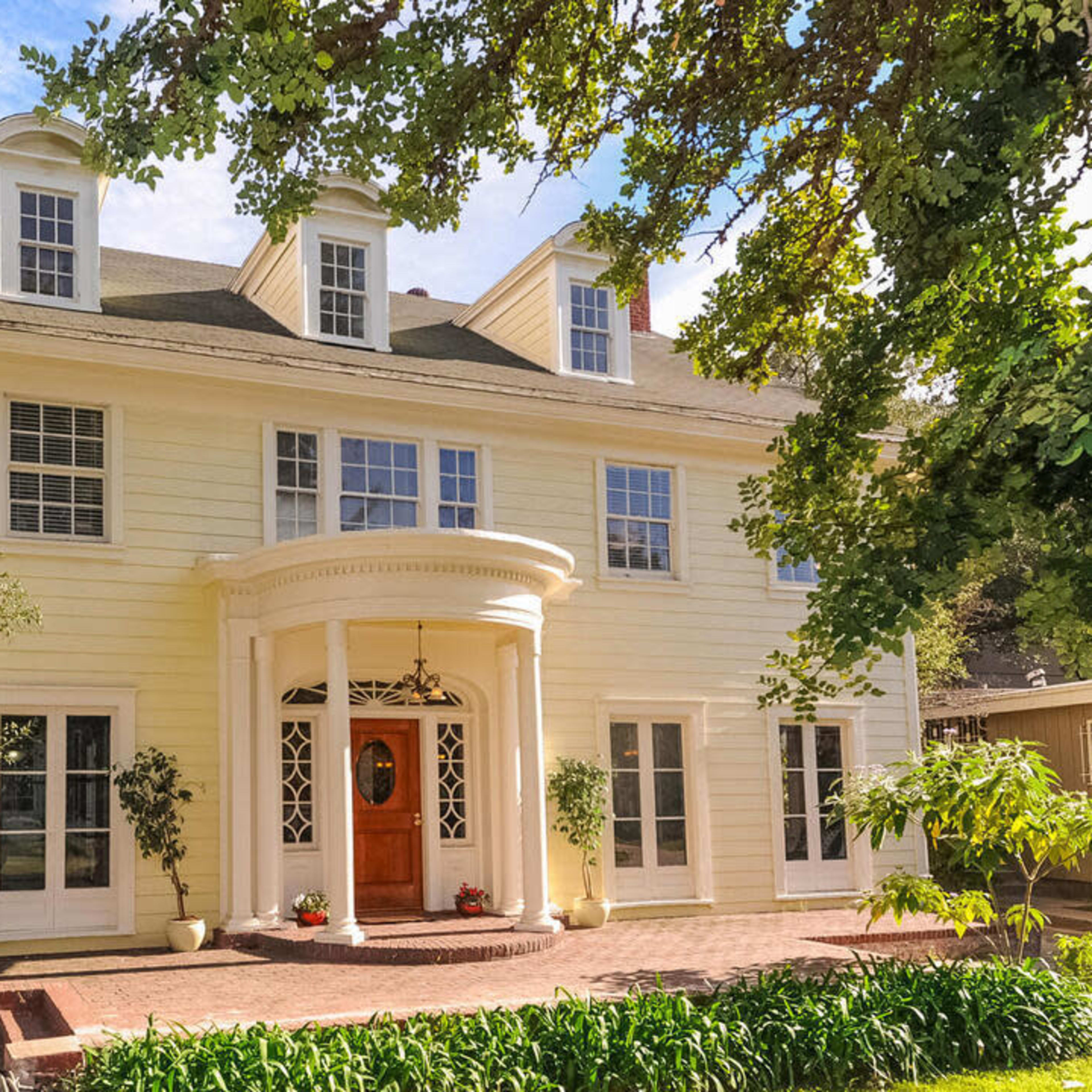 A large, two-story yellow house features a circular entryway and multiple windows surrounded by greenery.