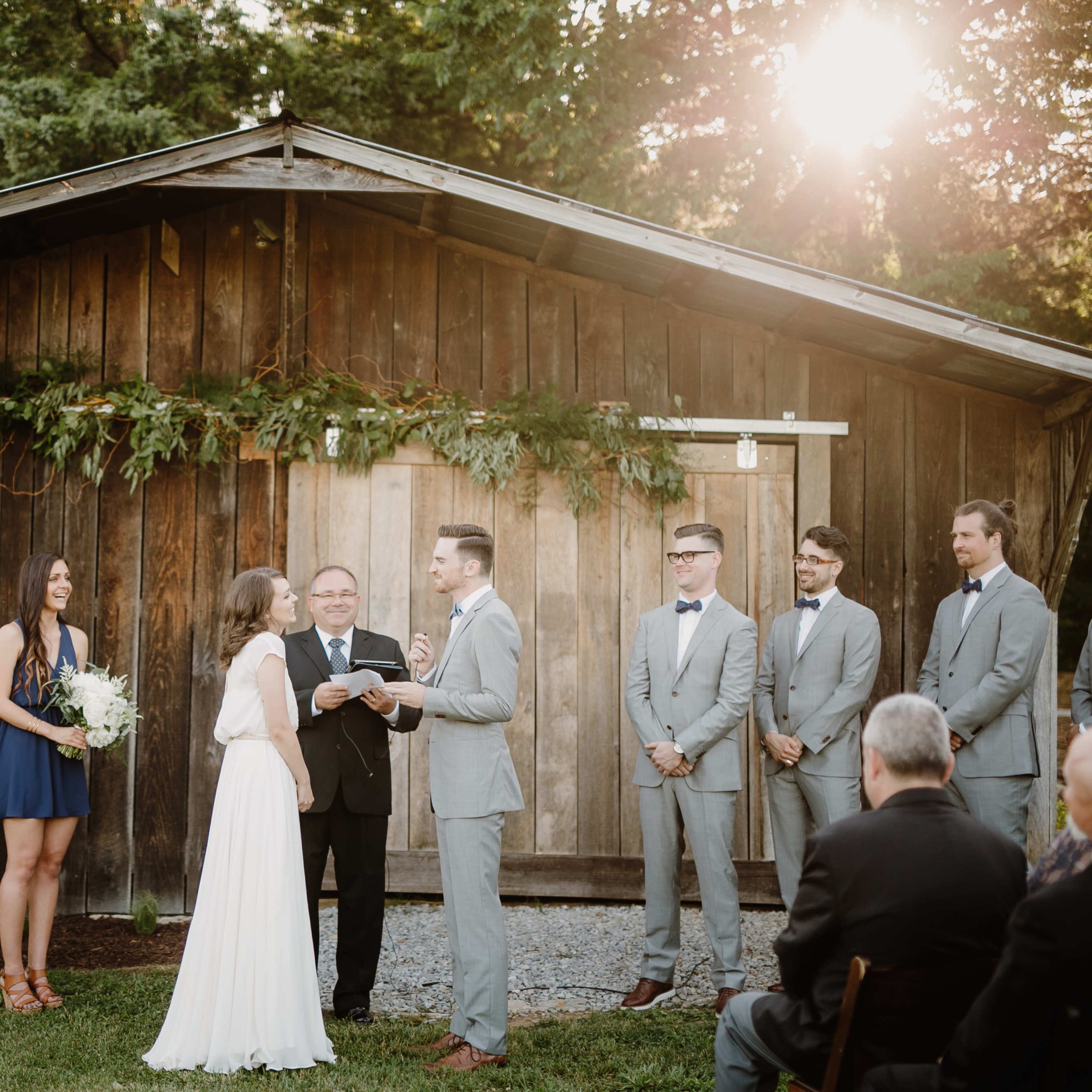 A couple exchanges vows during an outdoor wedding ceremony in front of a rustic wooden backdrop, surrounded by bridal party members and guests.