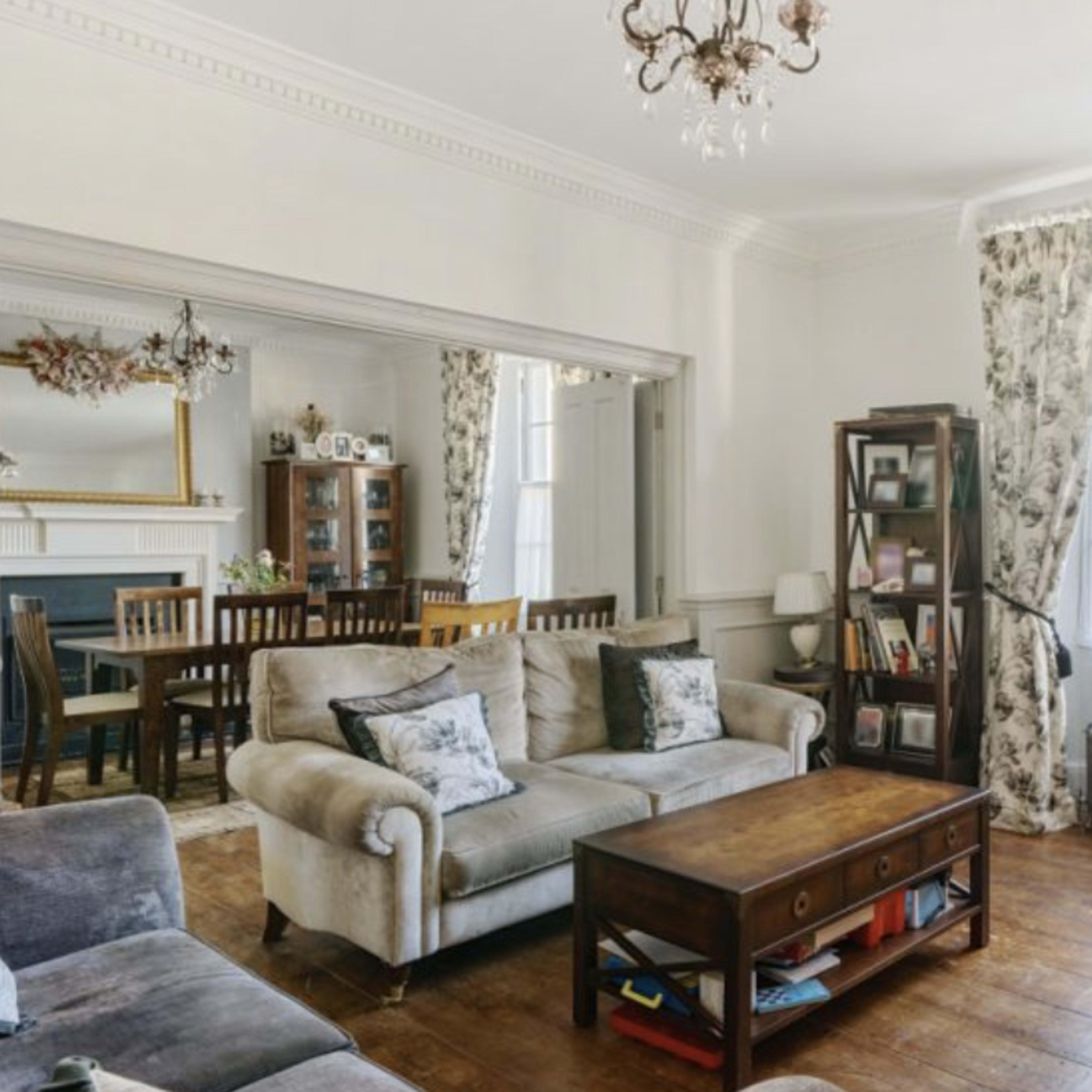 A spacious living room features a mix of light and dark furniture, a wooden coffee table, and large windows with floral curtains.
