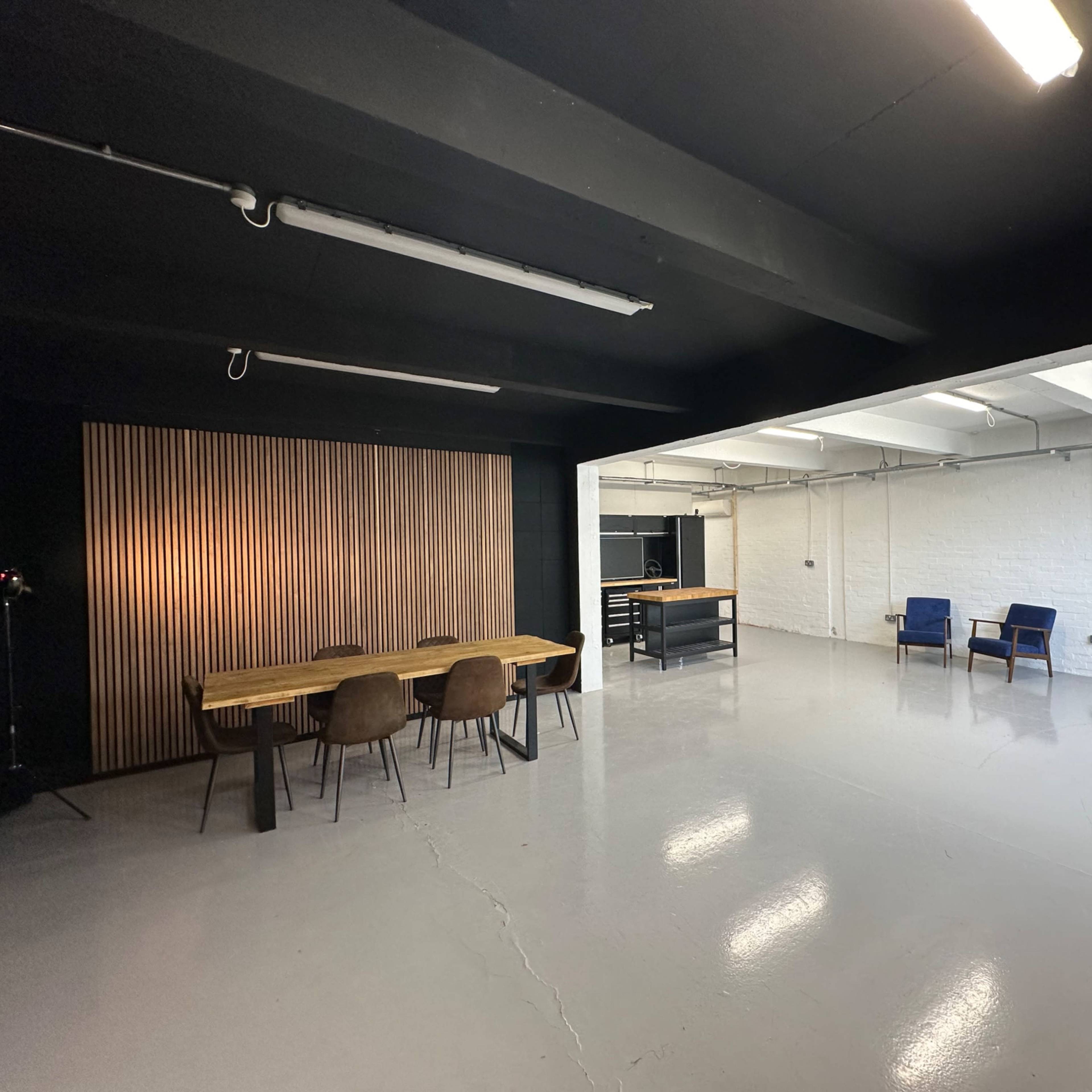 A spacious, well-lit room with a wooden dining table and chairs on one side, and two blue armchairs on the opposite side against a white wall.