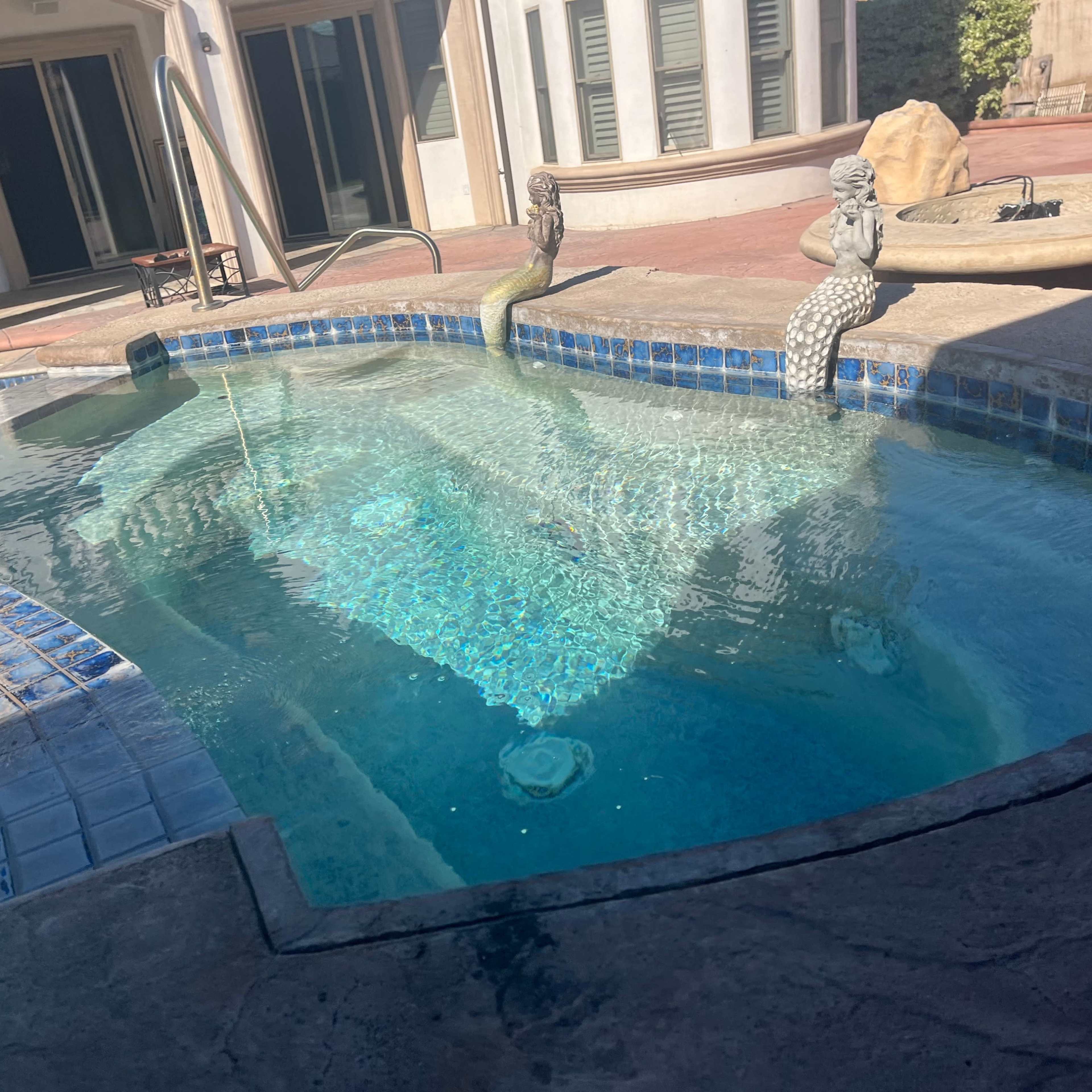 A decorative swimming pool features a stone mermaid sculpture and a fountain, surrounded by tiled edges and a patio area.