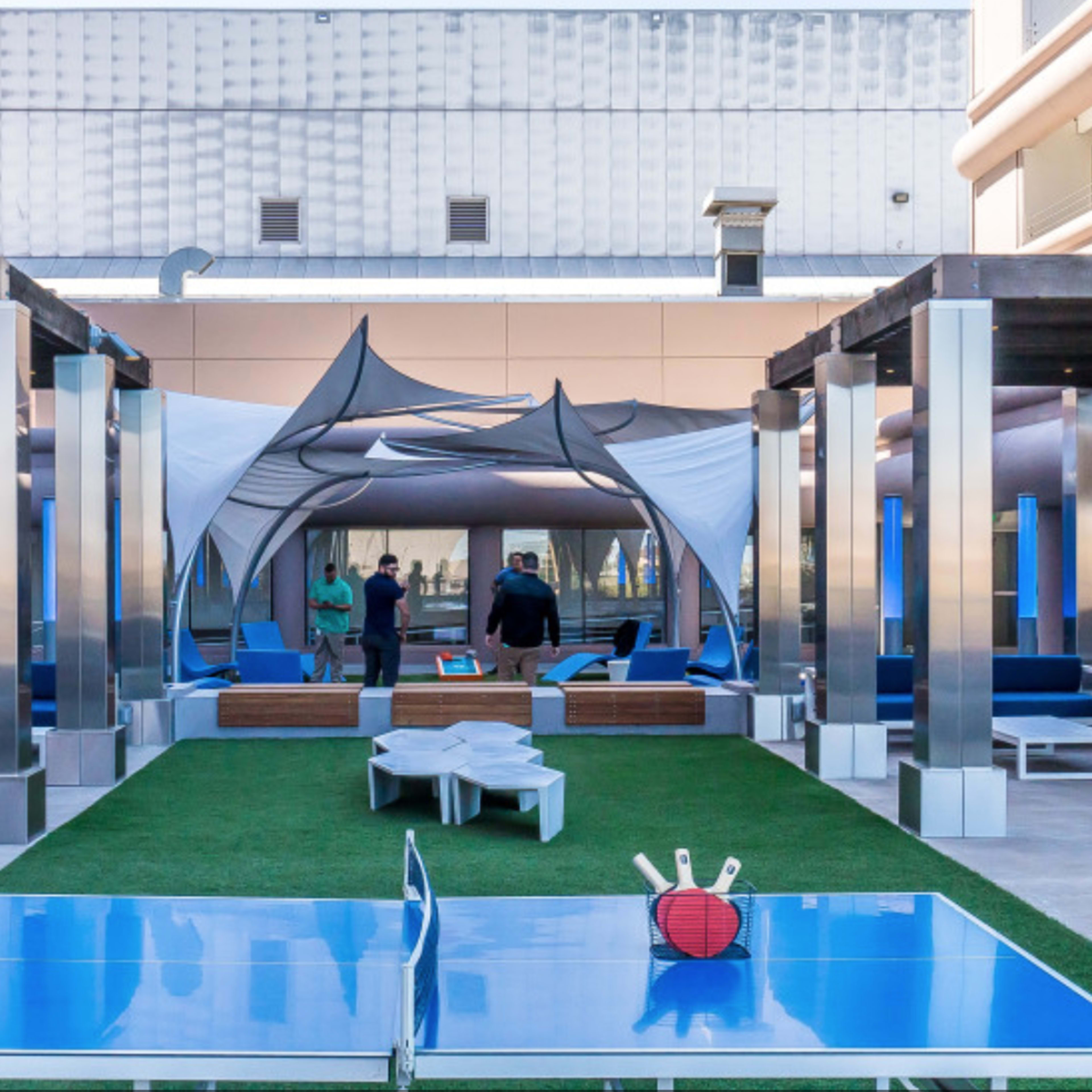 A rooftop lounge area with several seating arrangements, a large tent structure, and multiple ping pong tables on green turf.