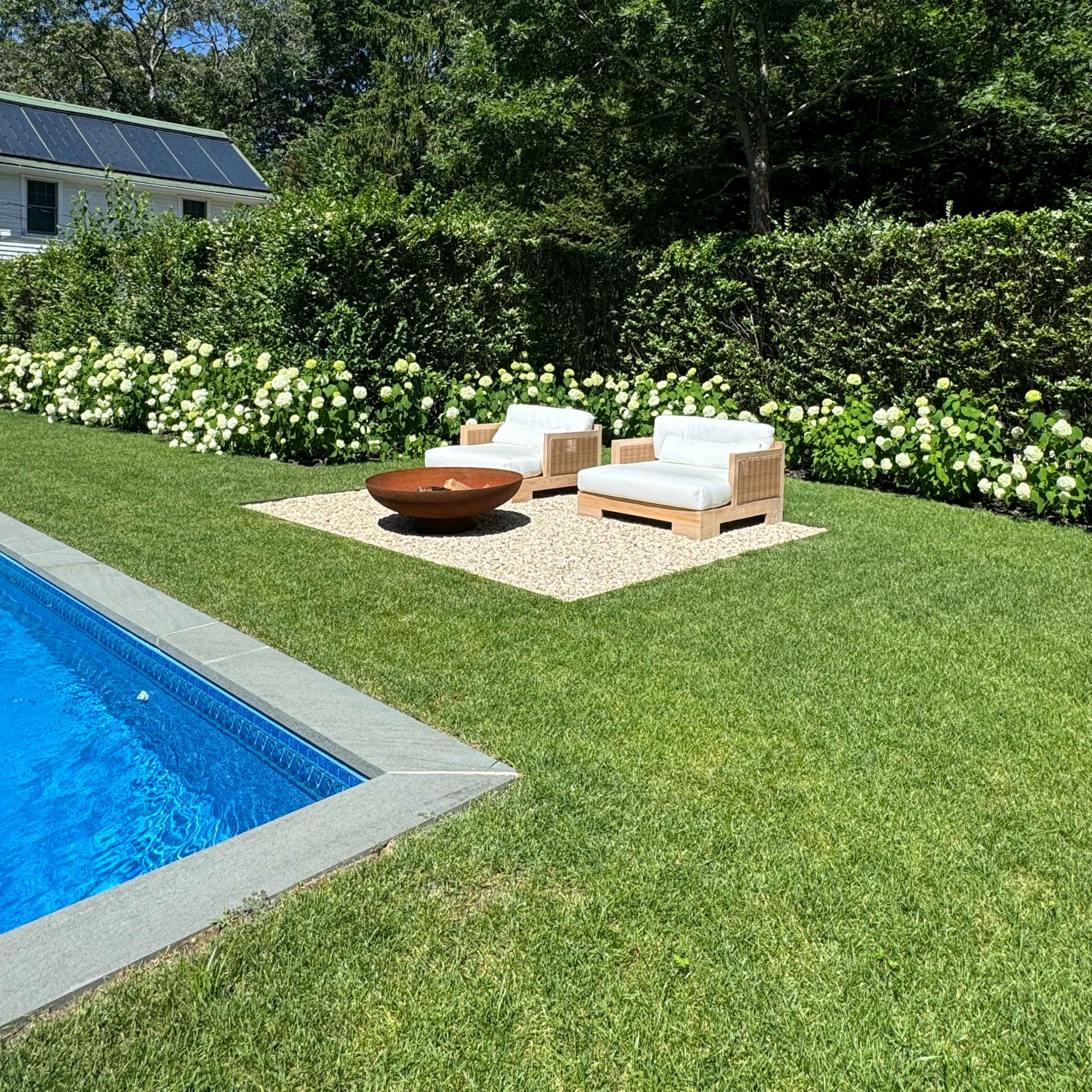 A poolside area with two white chairs and a round table surrounded by green grass and flowering bushes.