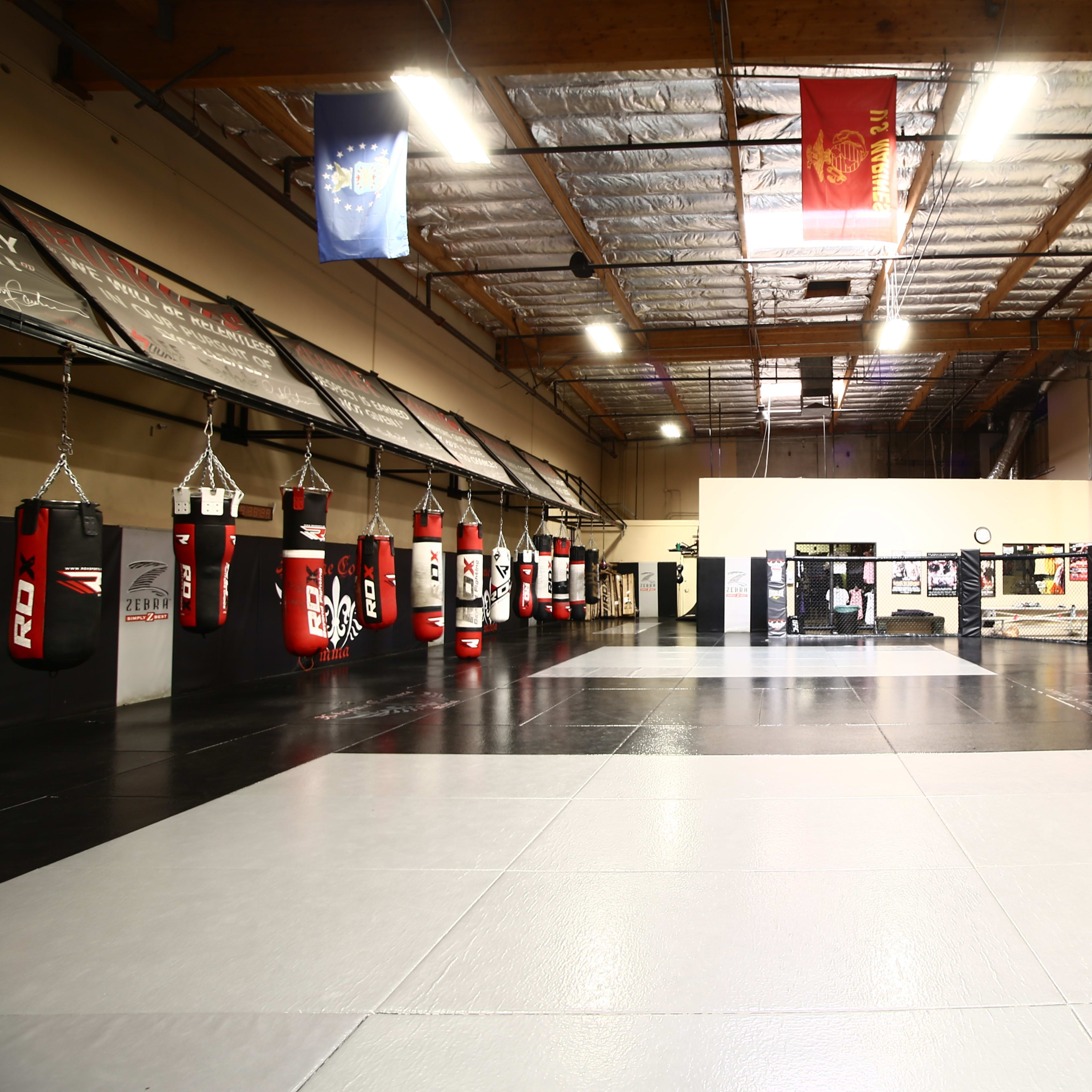 A spacious training facility for martial arts, featuring several hanging punching bags, a central mat area, and a cage surrounded by lockers and equipment.