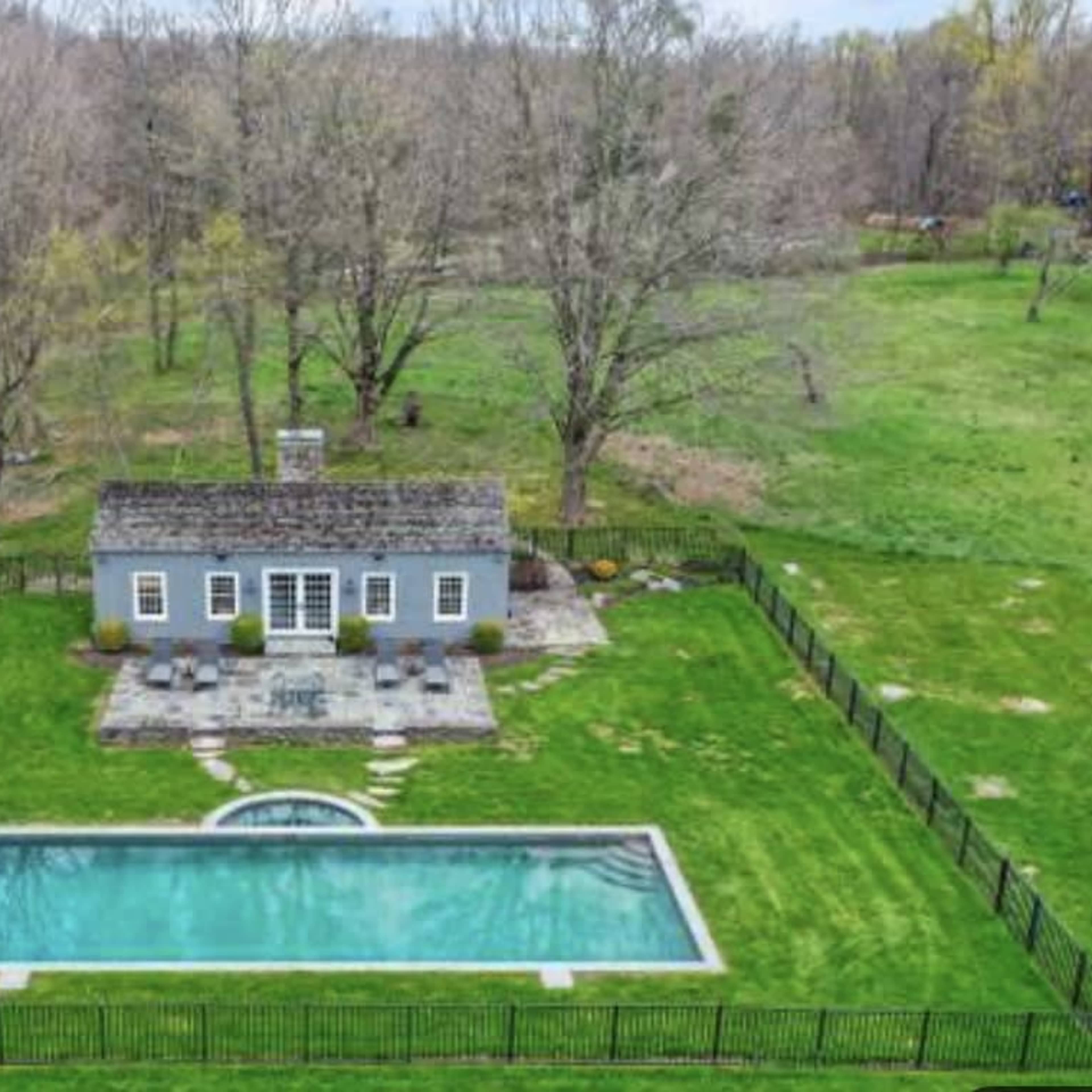 A backyard featuring a swimming pool, a small house, and a large green lawn surrounded by trees.