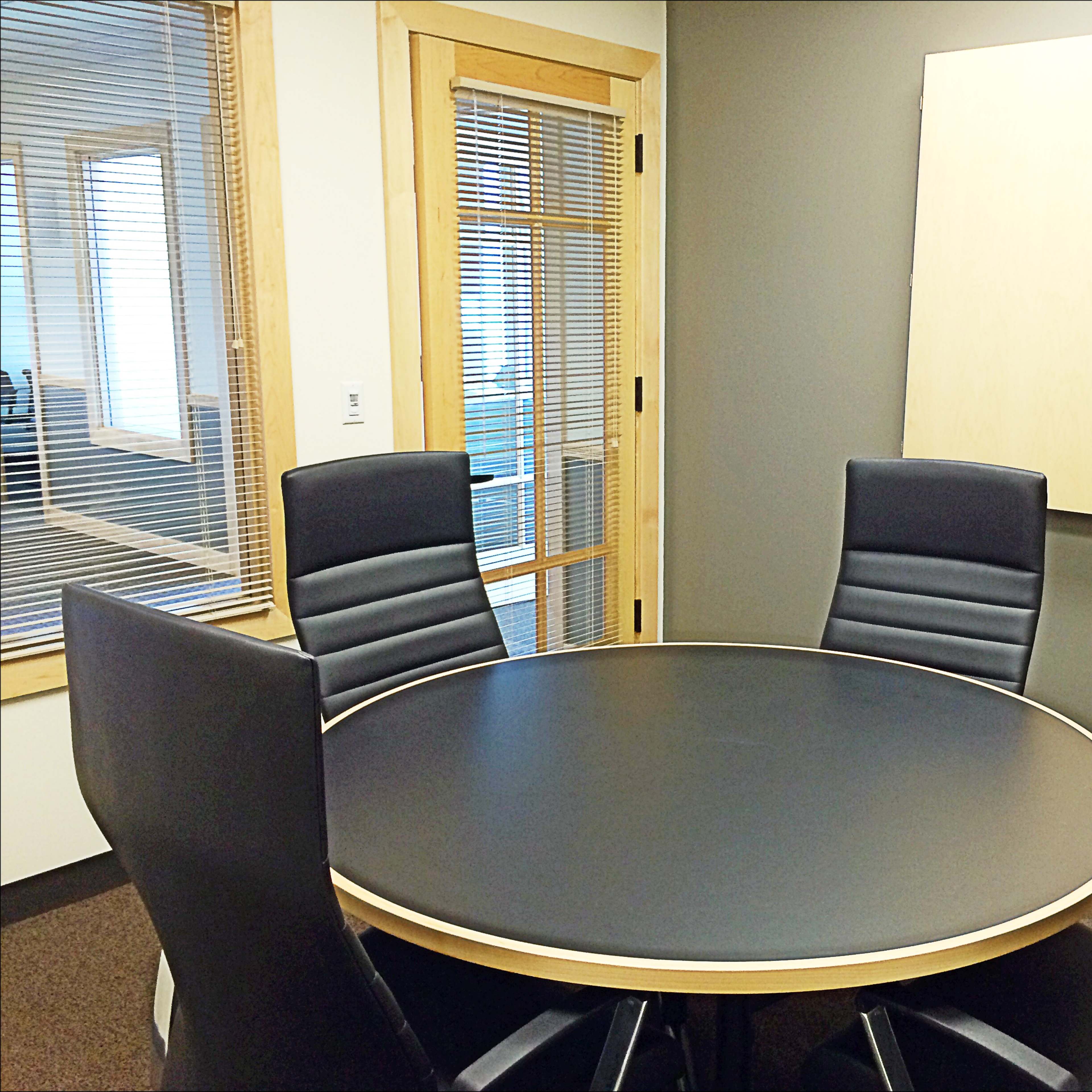 A circular conference table is set in a small meeting room with four black chairs and a wall-mounted cabinet.