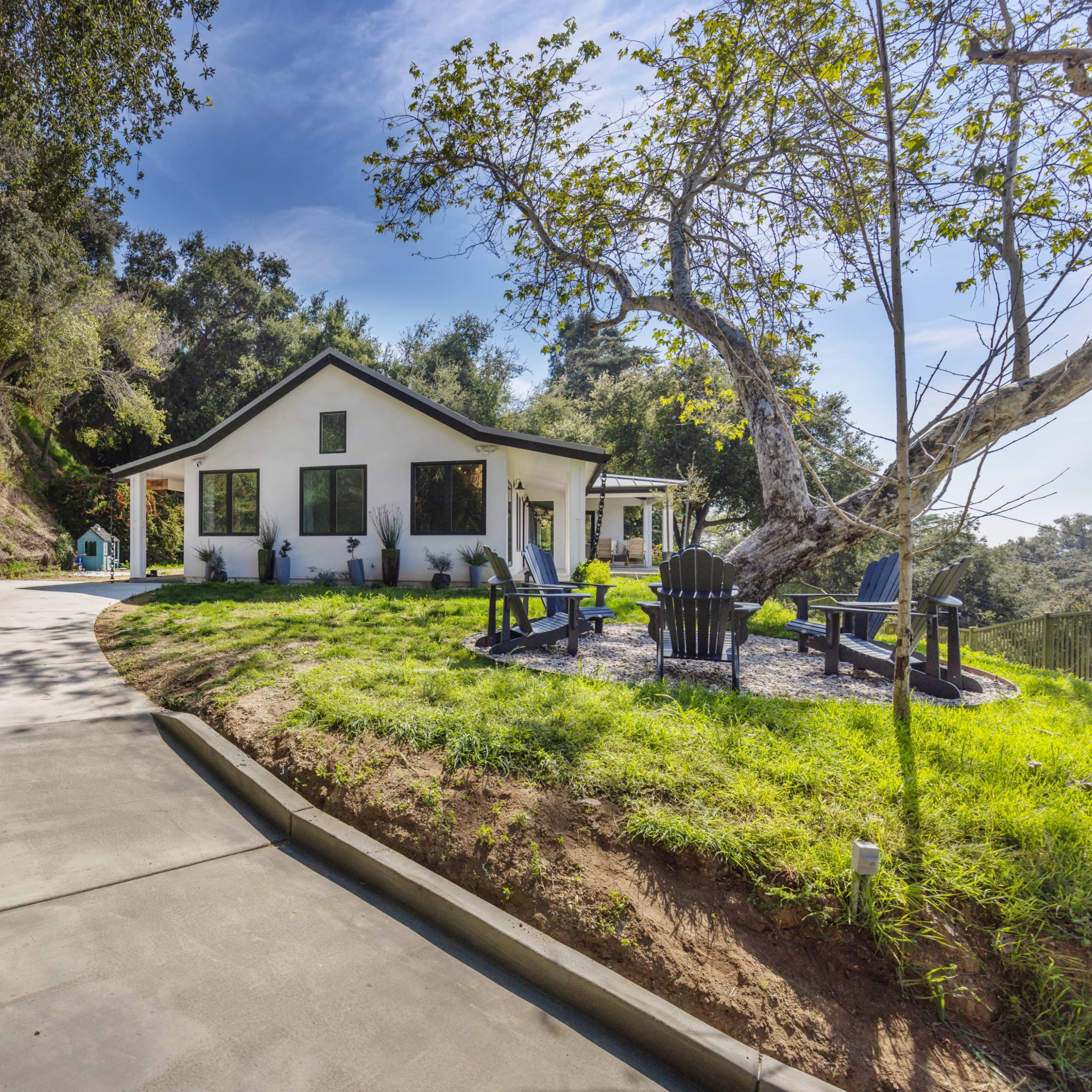 A modern house sits on a hillside, surrounded by trees, with a curved driveway leading to the front yard featuring outdoor seating.