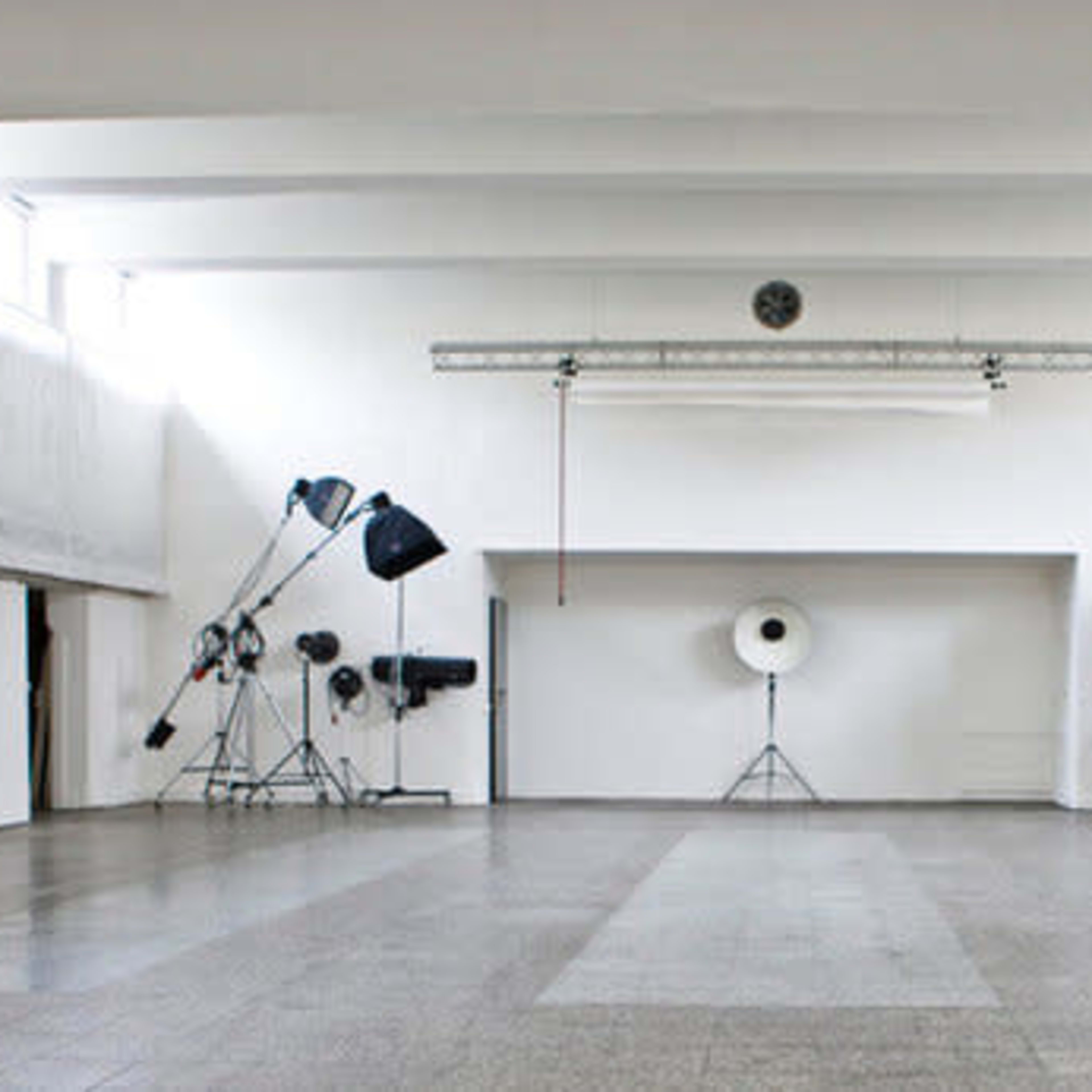 A large, empty studio space with white walls and various photography equipment, including light stands and large reflectors, arranged in the corners.