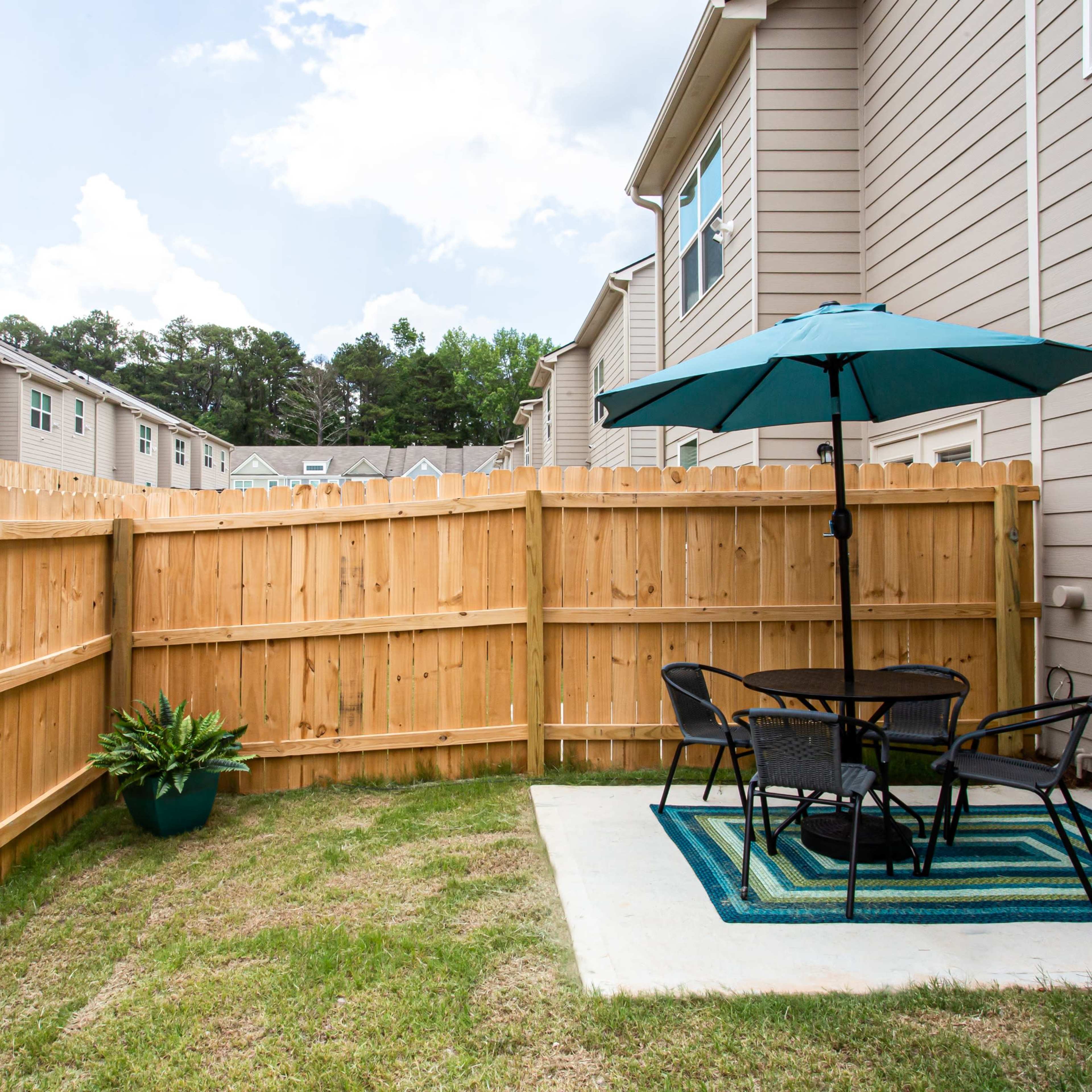 A small patio area features a table and chairs under a blue umbrella, surrounded by a wooden fence and grass.