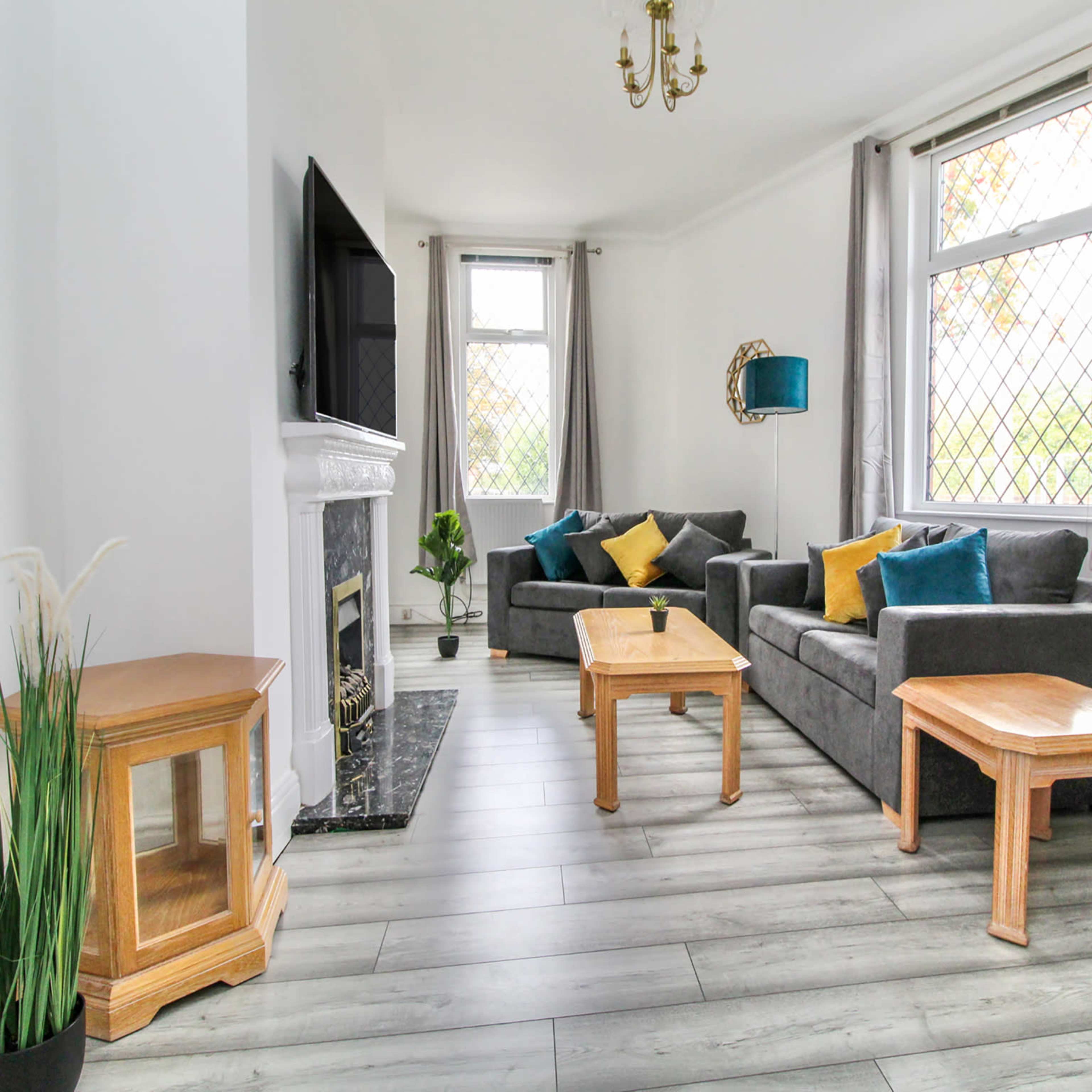 A tidy living room features two gray sofas with colorful throw pillows, wooden tables, and large windows allowing natural light.