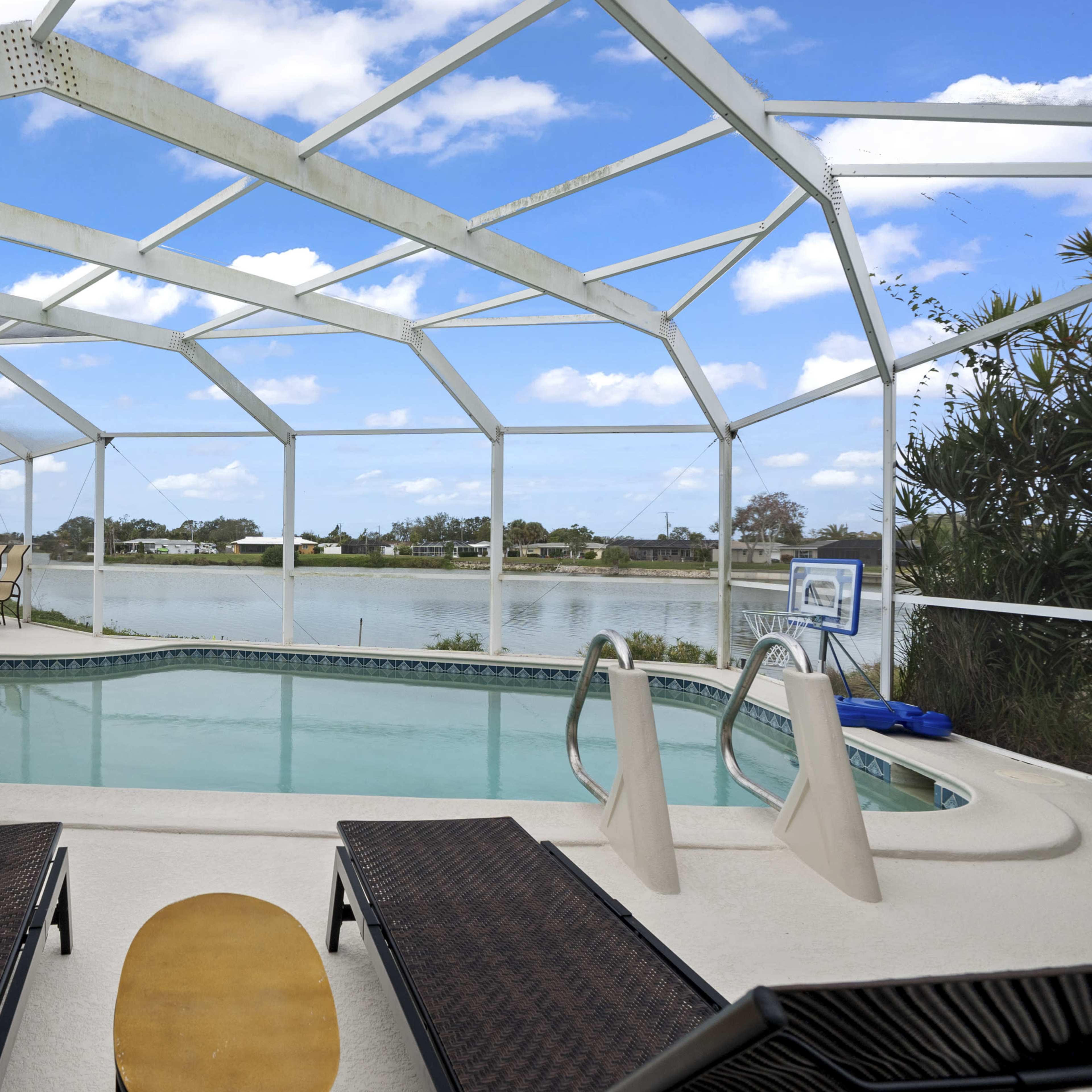A screened-in patio features a swimming pool with two lounge chairs and a view of a lake and blue sky.