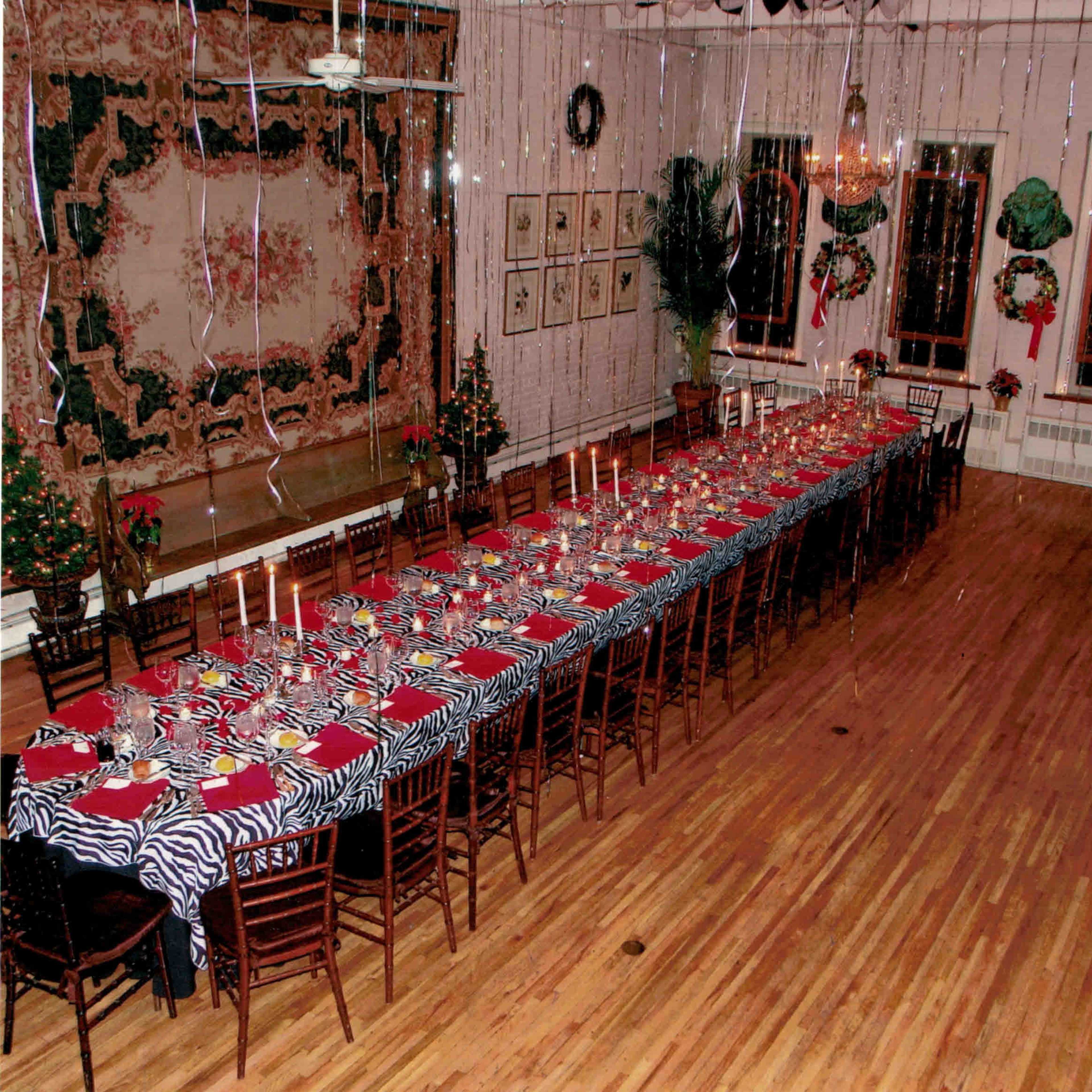 A long dining table with a zebra-patterned tablecloth is set with red plates and candles, located in a decorated room featuring festive hanging decorations and a chandelier.