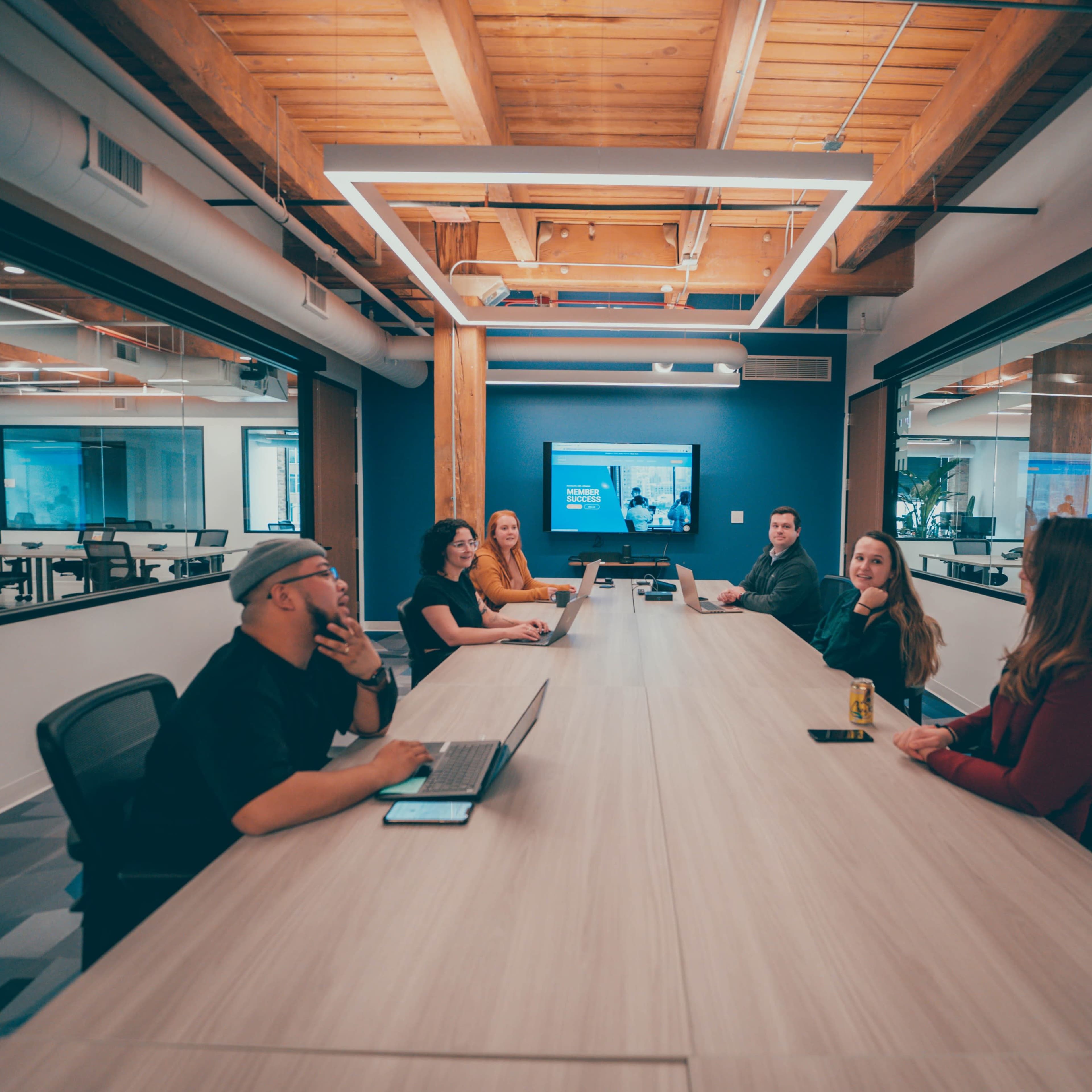 A group of six people sit around a long, wooden conference table in a modern office meeting room.