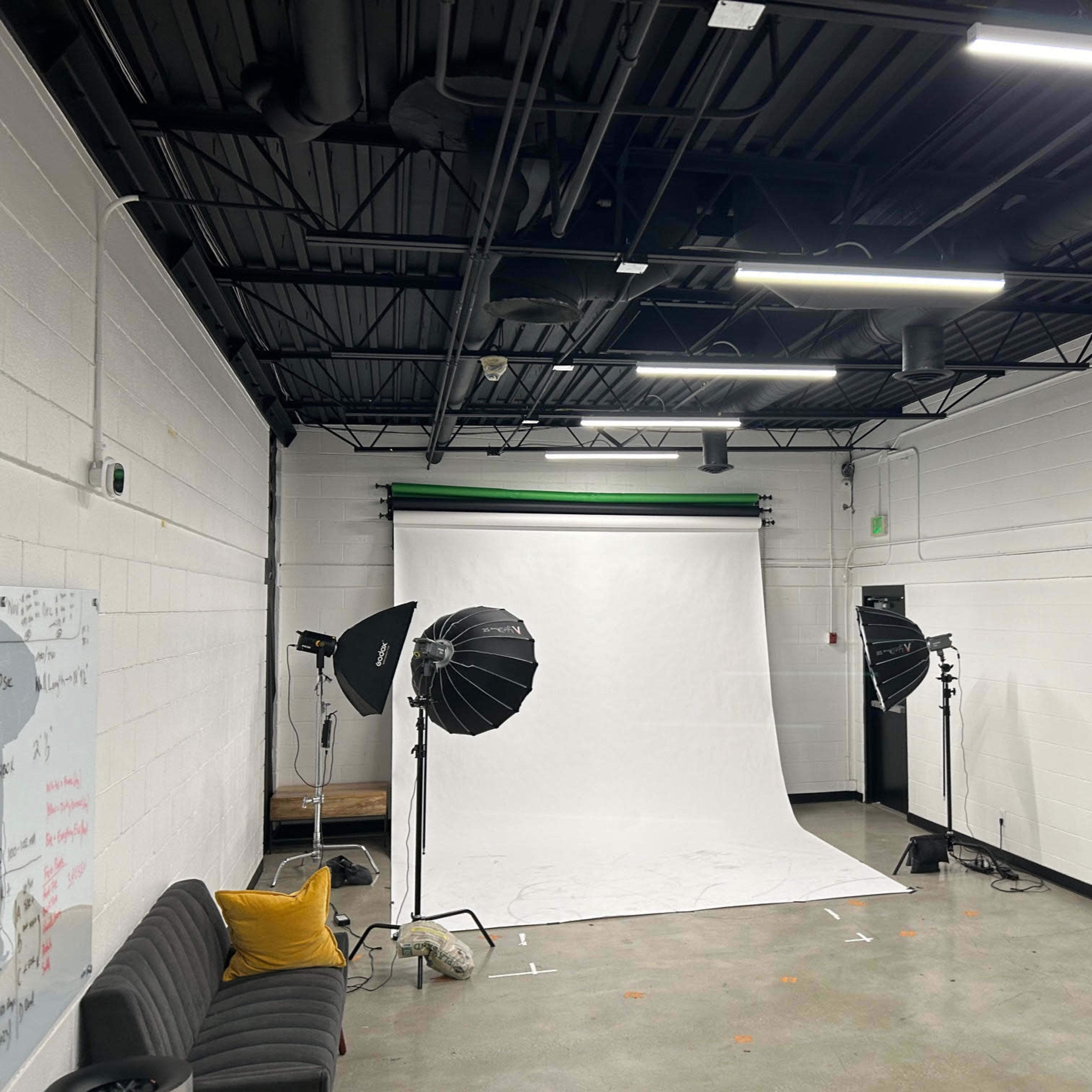 A photography studio with a white backdrop, lighting equipment, a black sofa, and two computer monitors on a desk.