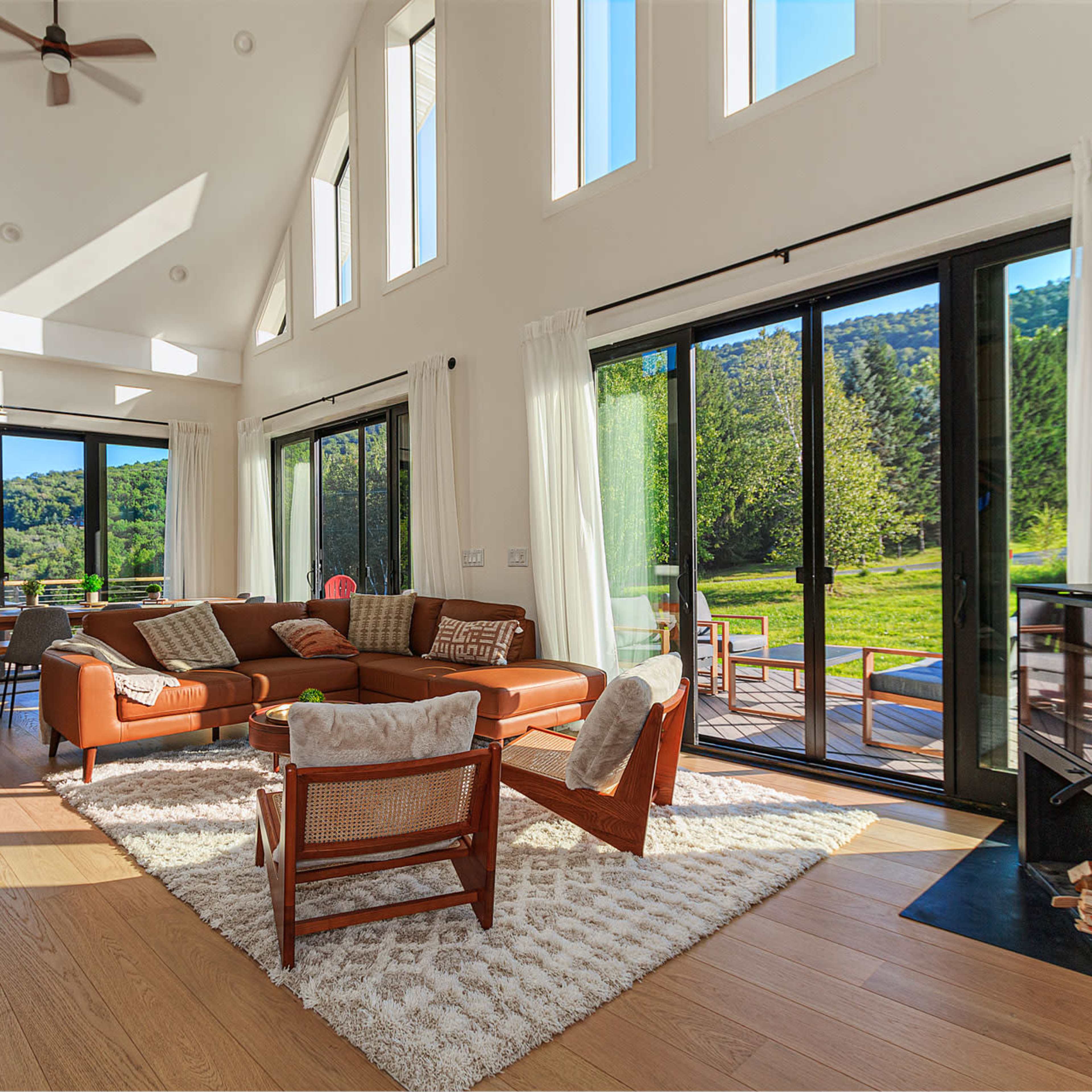 The interior of a modern living room features large windows, a brown sofa, wooden flooring, and a view of lush greenery outside.