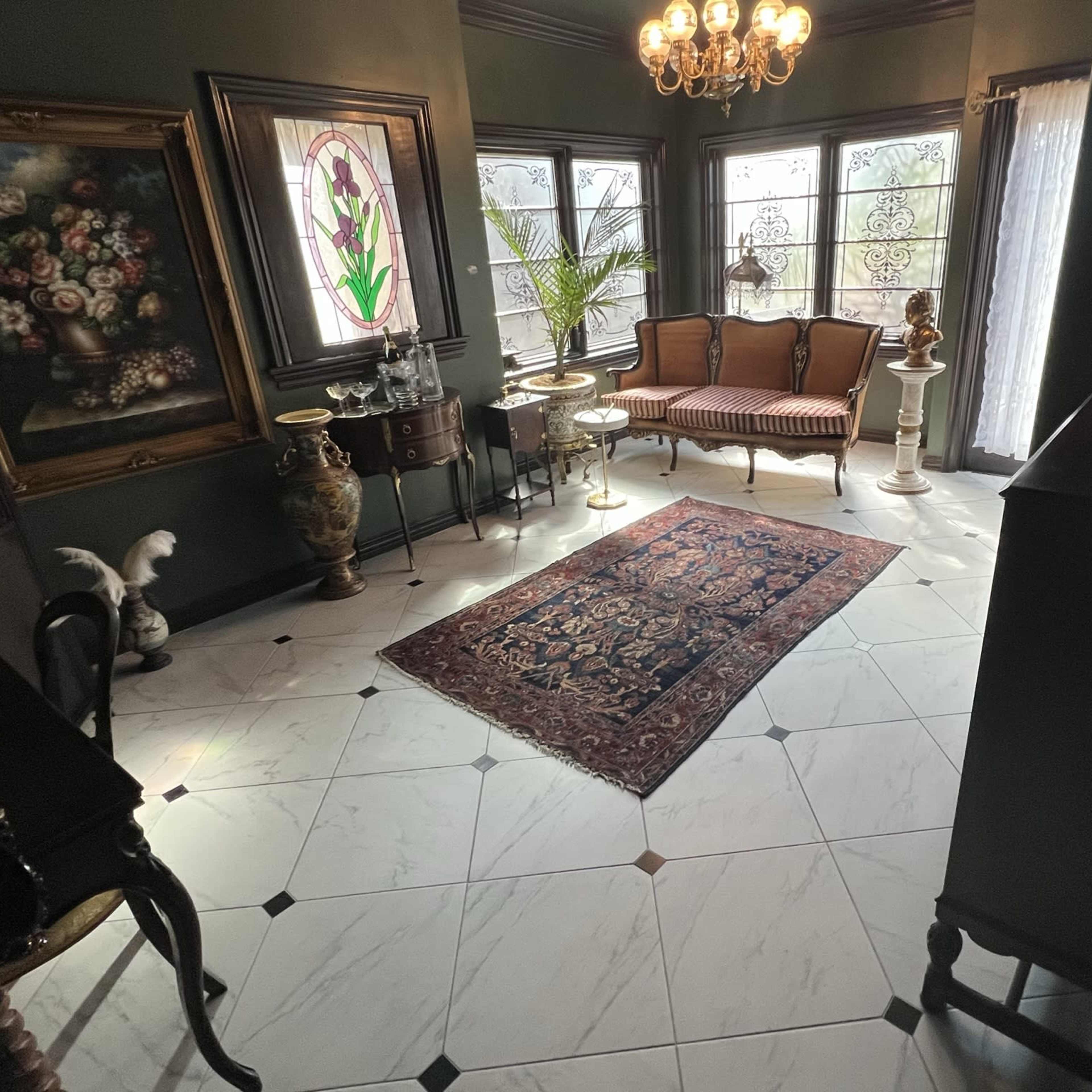 An elegantly furnished room with a patterned rug, vintage furniture, and decorative art on the walls, illuminated by natural light.