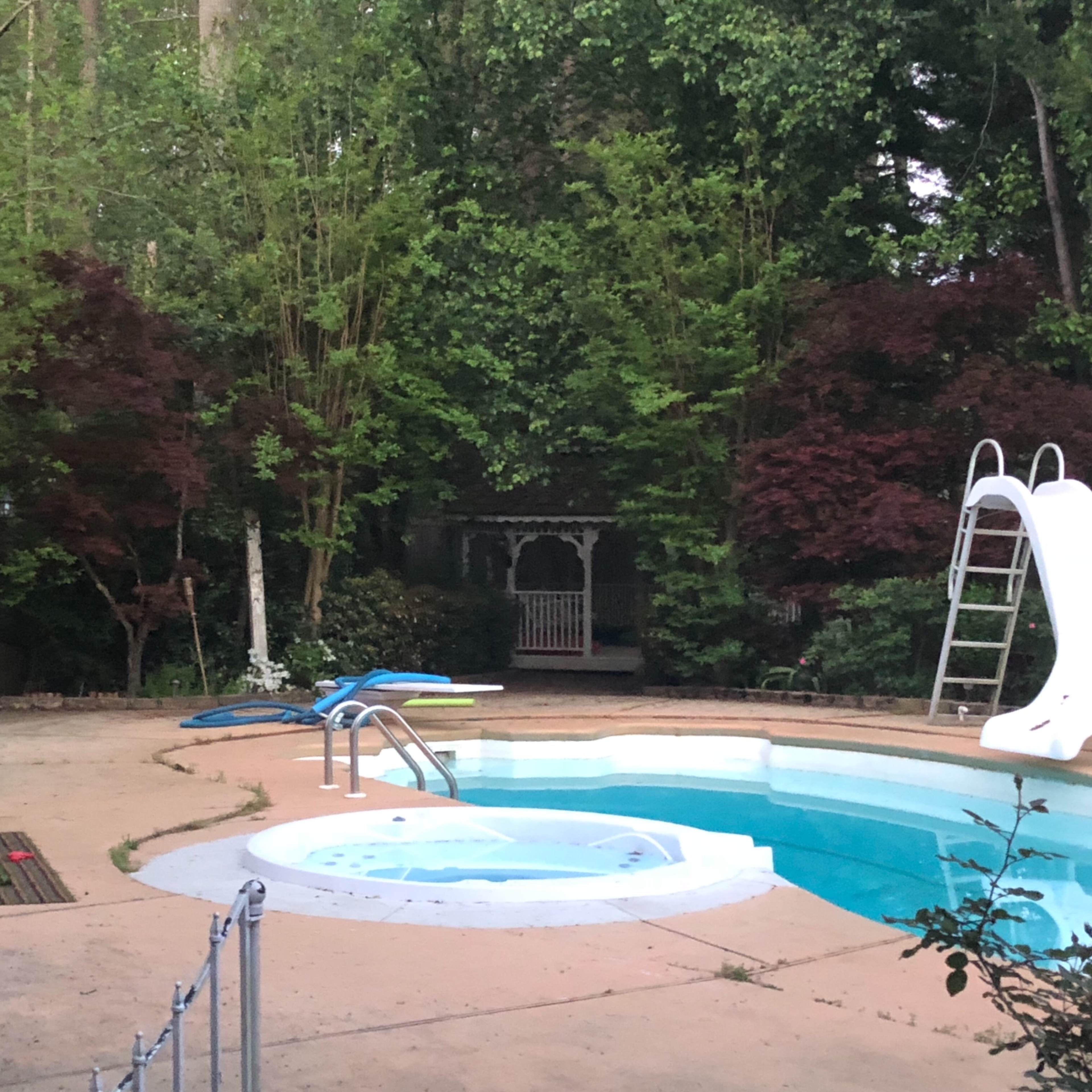 A swimming pool with a slide and a hot tub is surrounded by greenery and features a gazebo in the background.