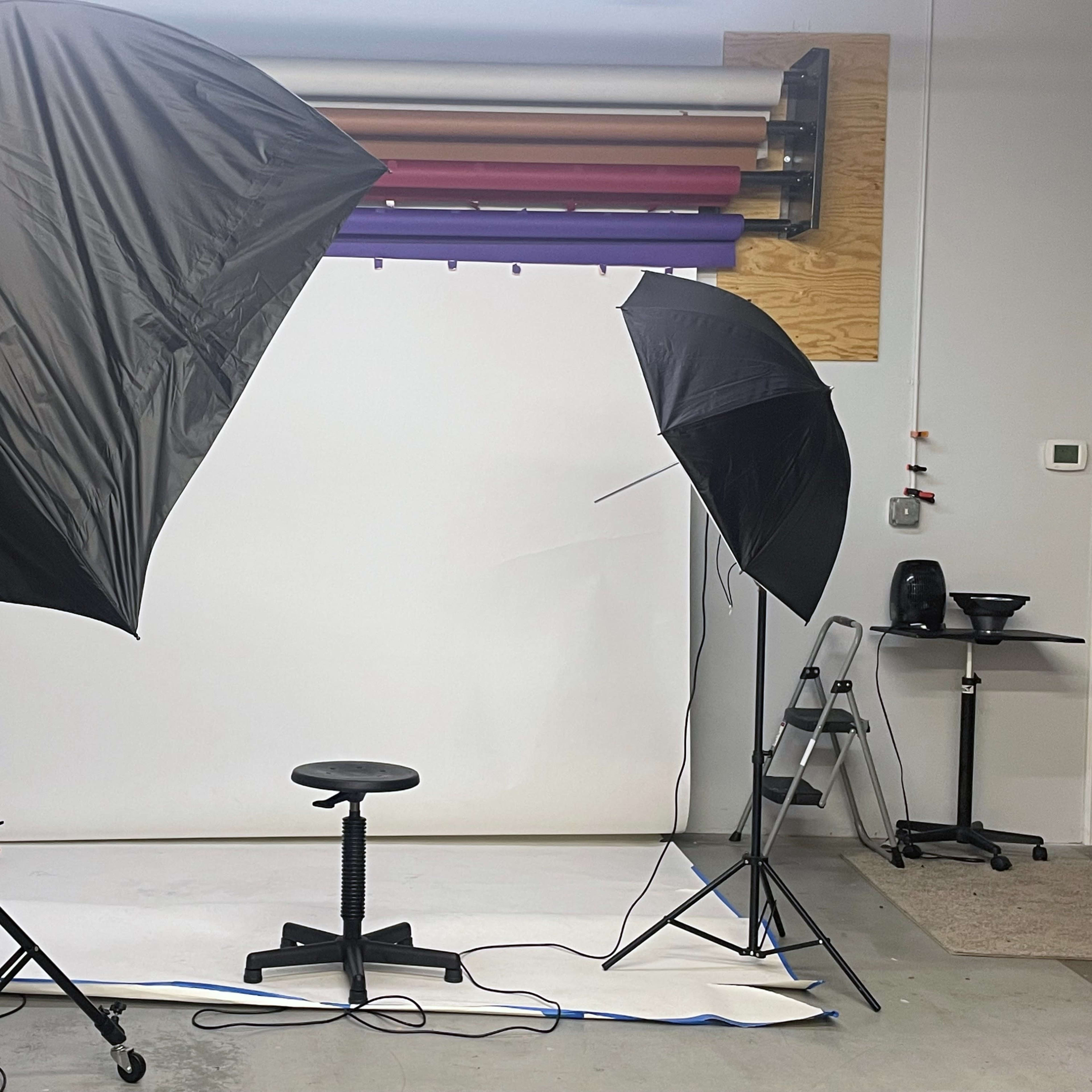 A photography studio setup with two umbrellas, a stool in the center, and various backgrounds hung on the wall.