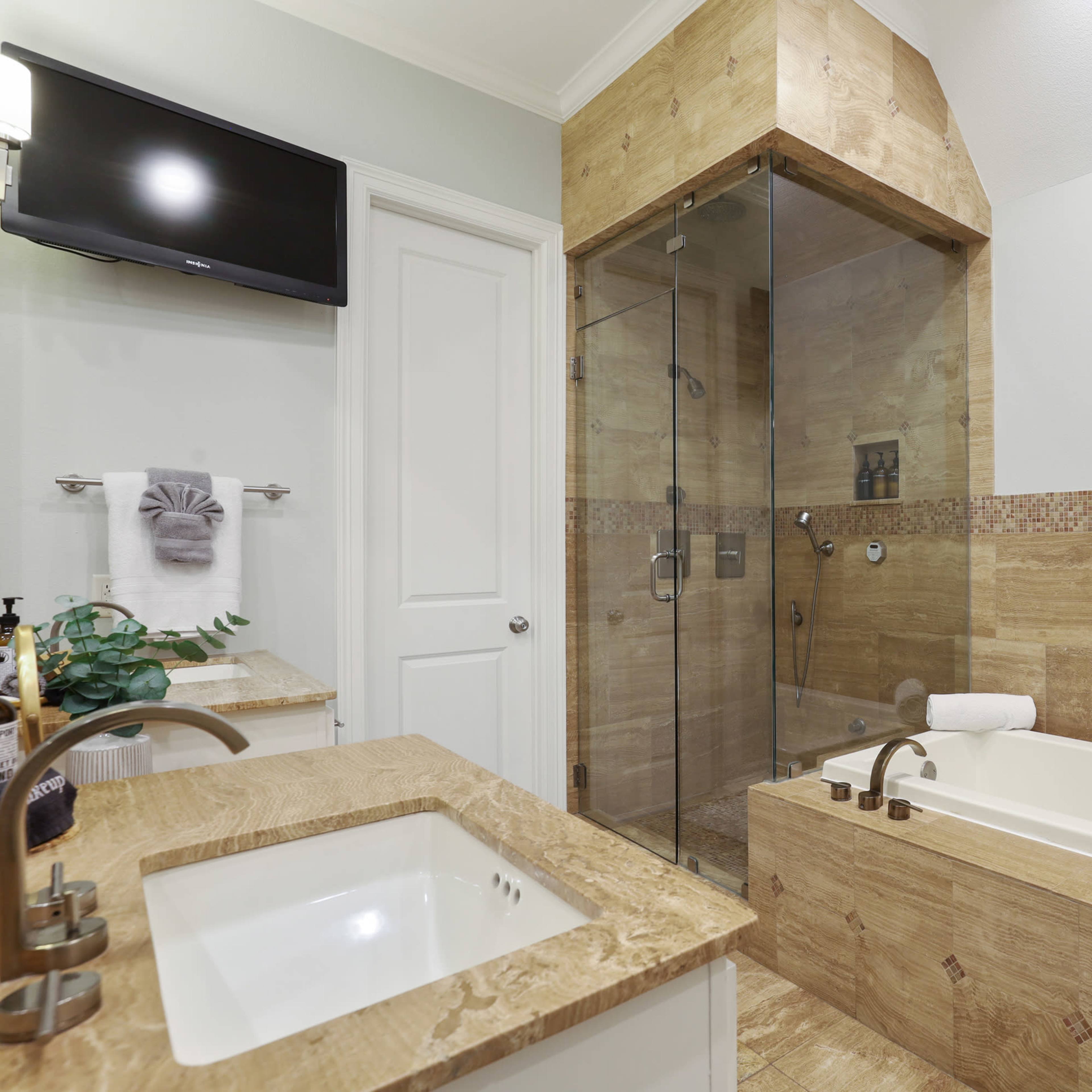 A bathroom featuring a glass shower enclosure, a freestanding bathtub, a double sink vanity with a stone countertop, and a television mounted on the wall.
