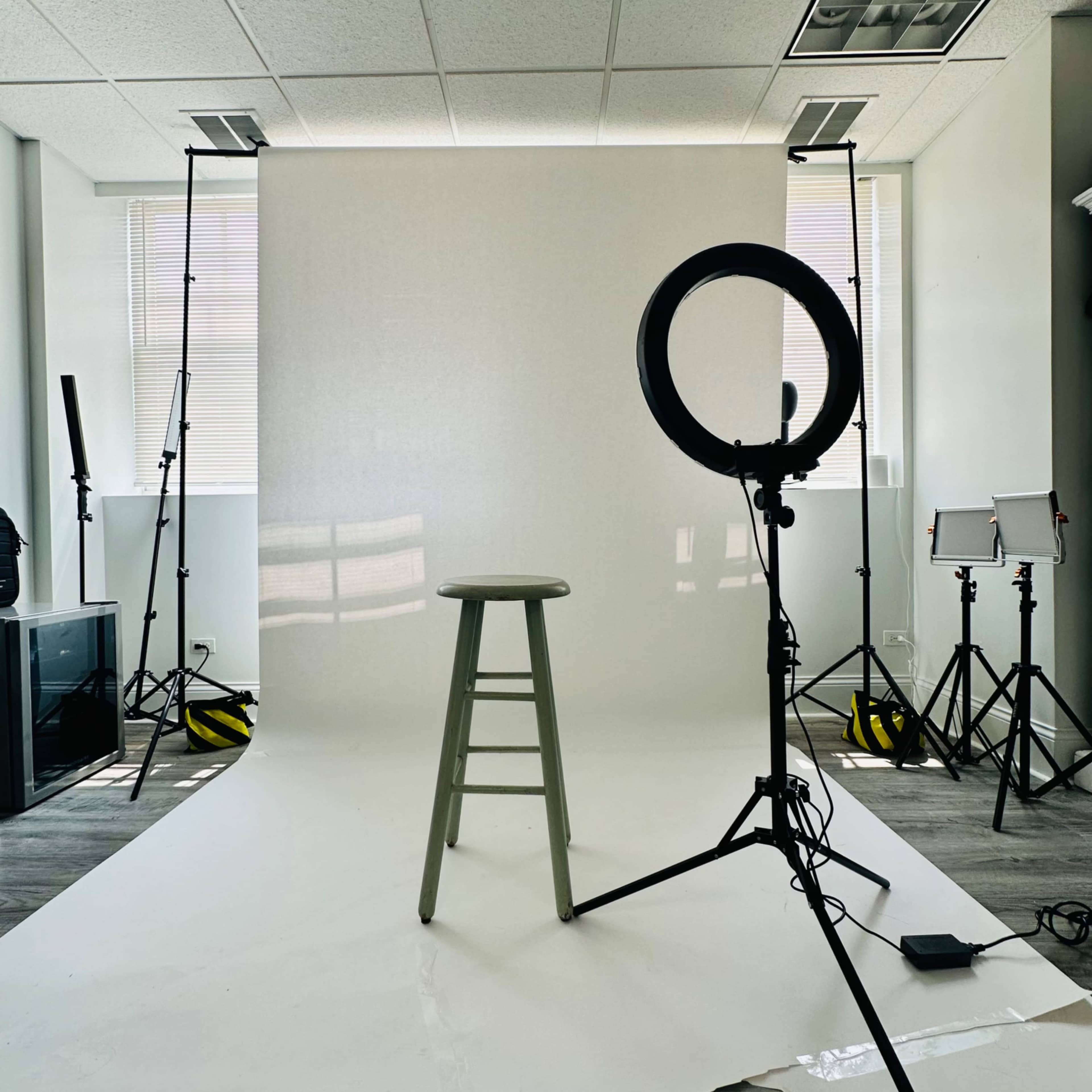 A photo studio setup with a gray stool placed in front of a white backdrop, surrounded by lights and a ring light.