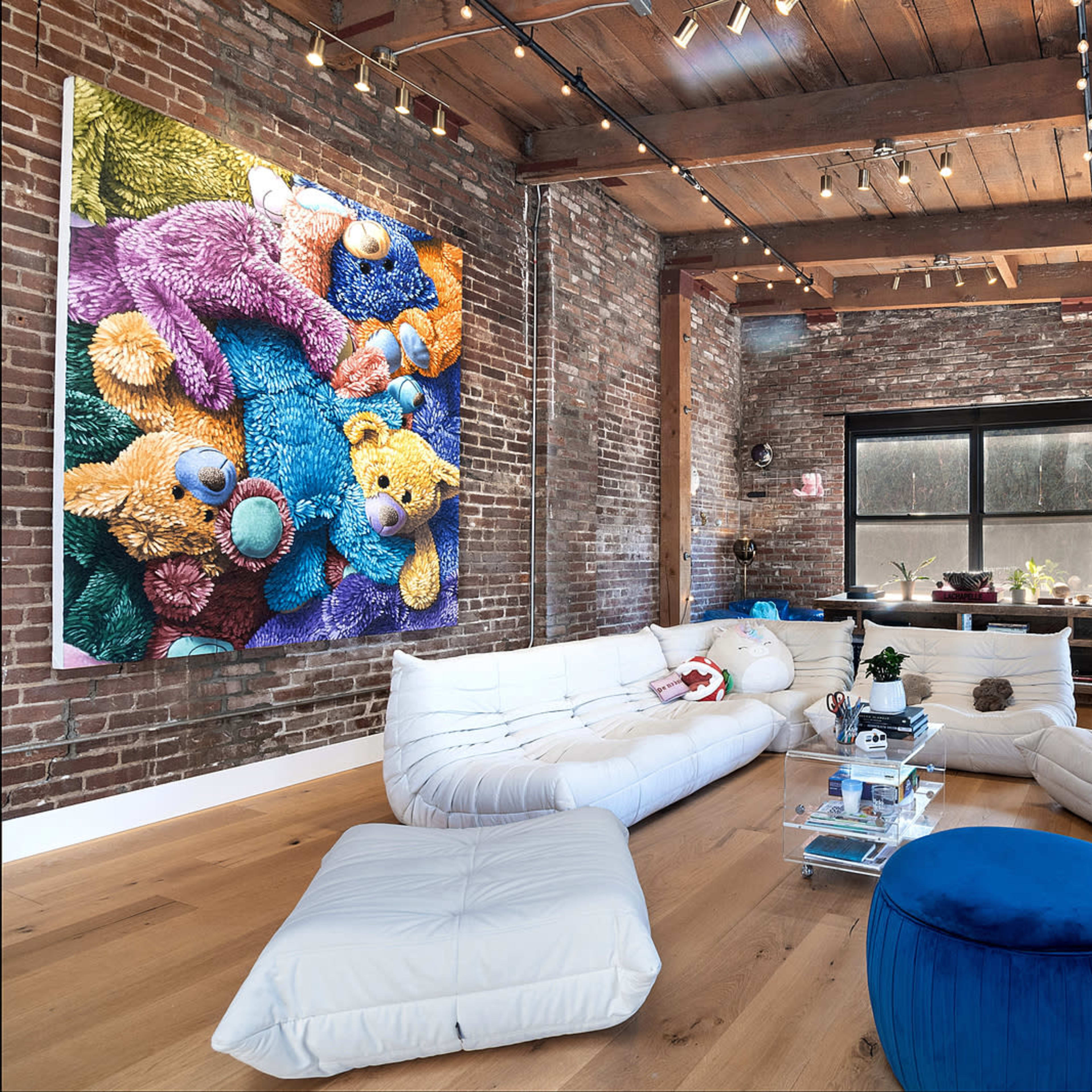 A spacious living area with exposed brick walls, a large colorful bear-themed artwork, and various seating options, including white lounge cushions and a blue ottoman.