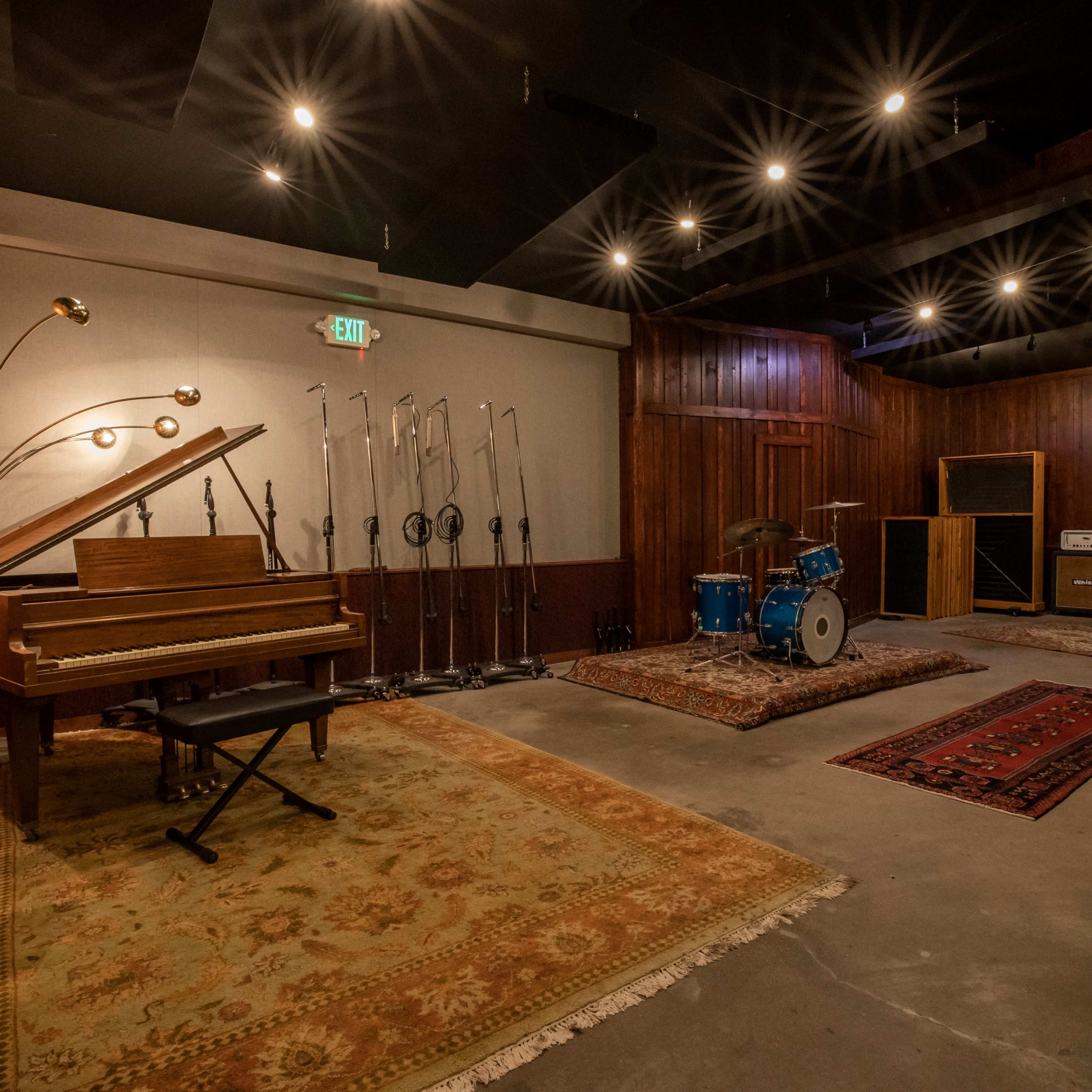 A music studio featuring a piano, a drum set, and various microphones arranged on stands in a spacious room with wooden walls and rugs.