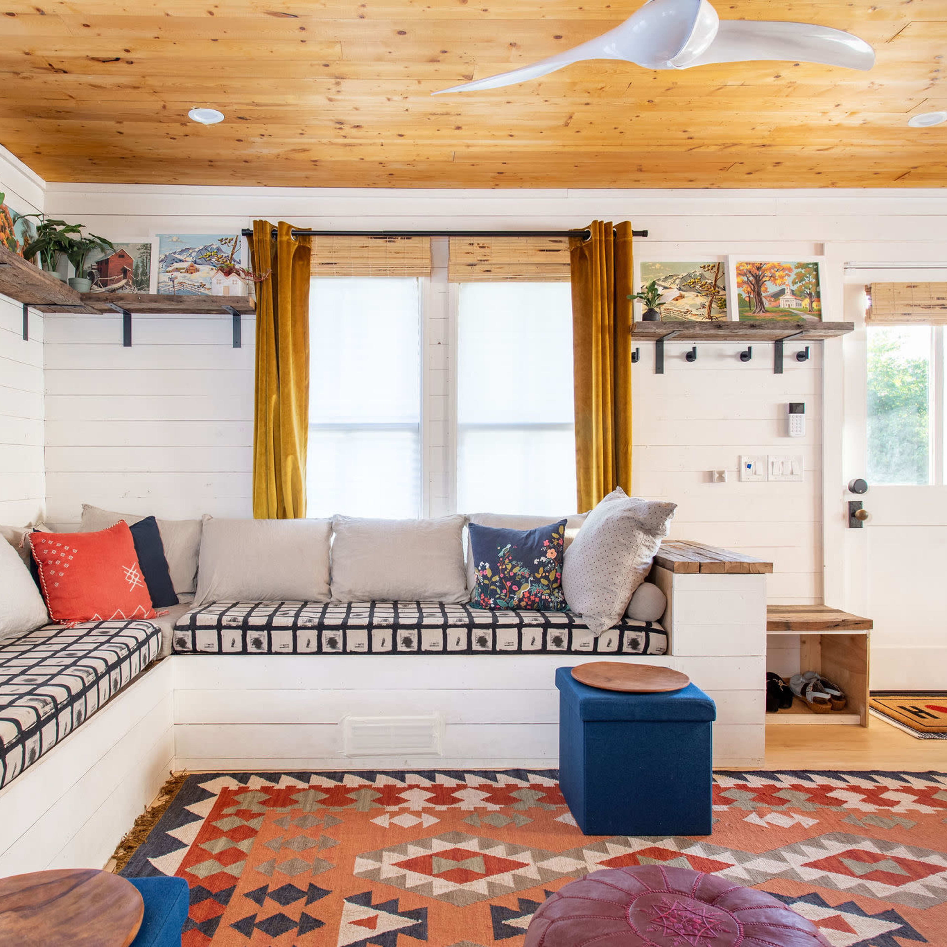 A cozy living room with a built-in seating area, wooden ceiling, decorative rugs, and bright curtains.