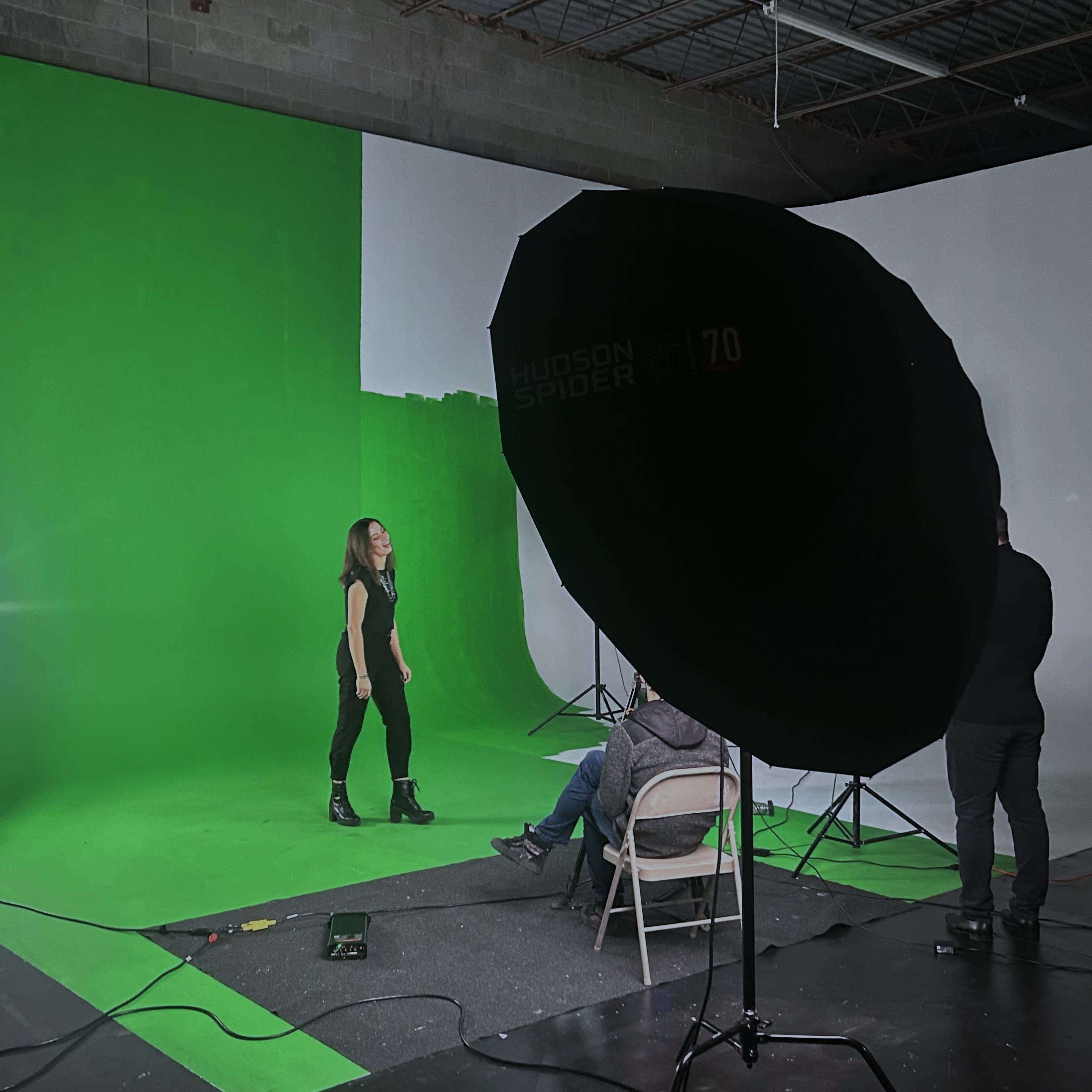 A model stands on a green backdrop while photographers adjust lighting equipment in a studio.