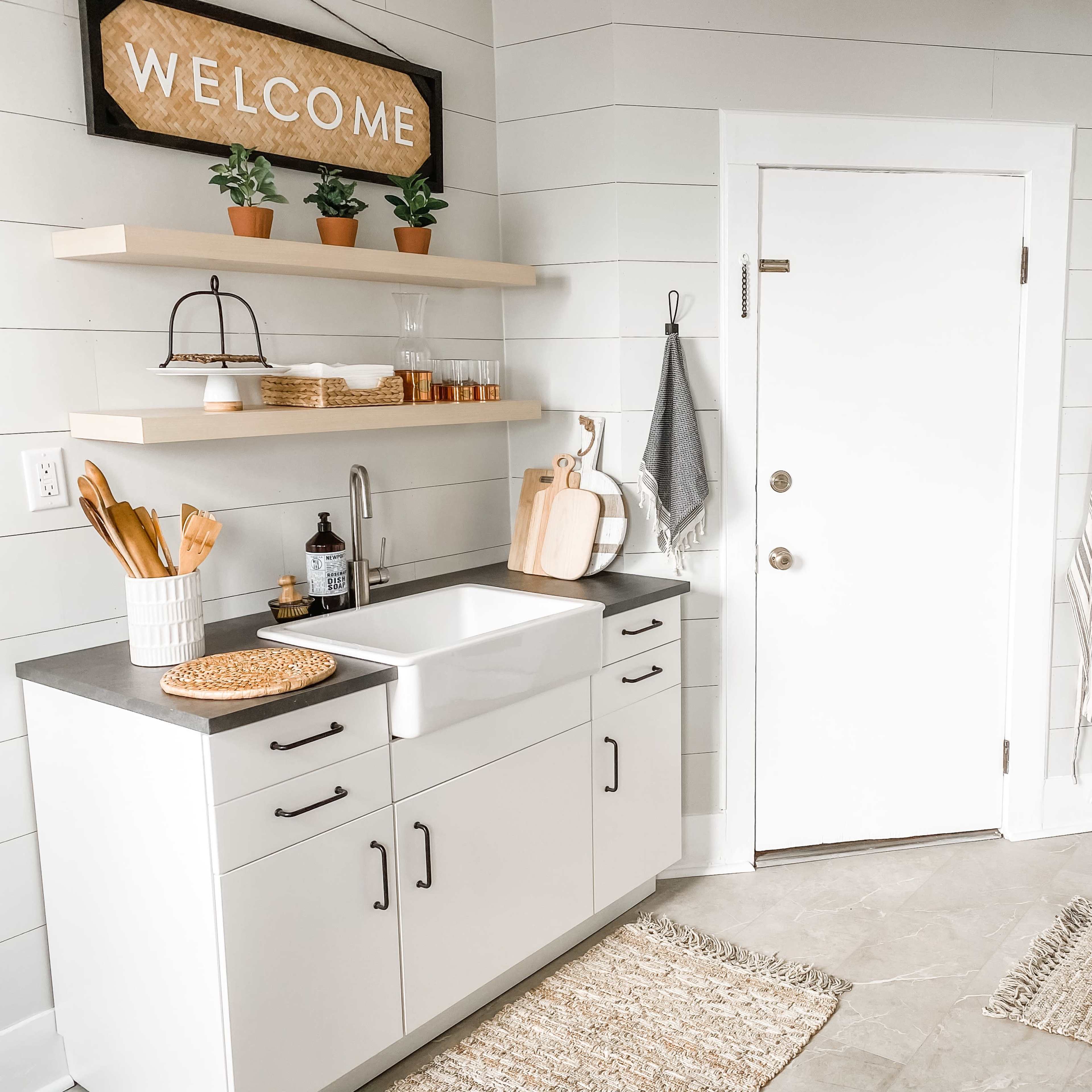 A modern kitchen features a white farmhouse sink, open shelving with potted plants, and a welcoming sign on the wall.