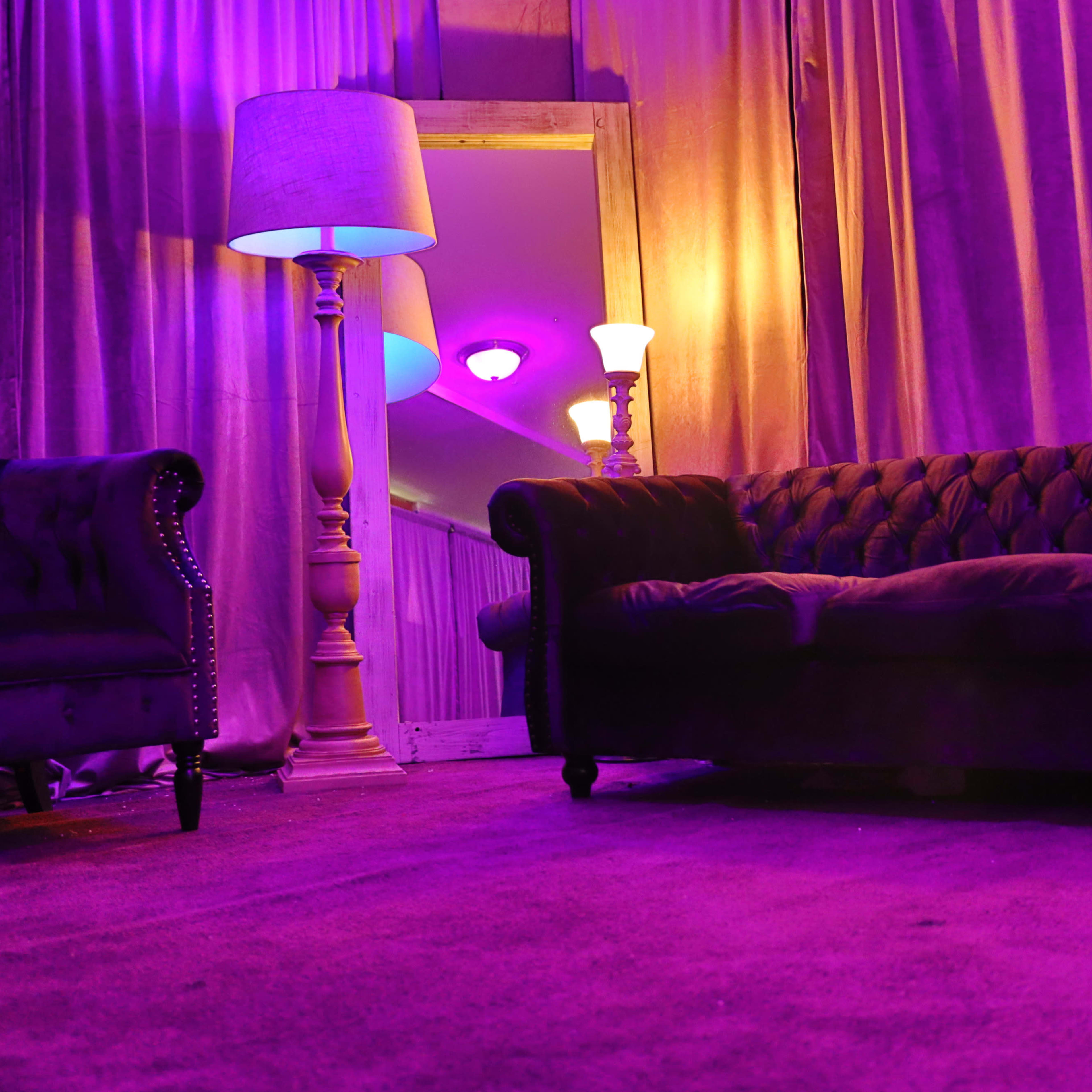 A velvet sofa and armchair are positioned in a room with purple curtains and ambient lighting from lamps.