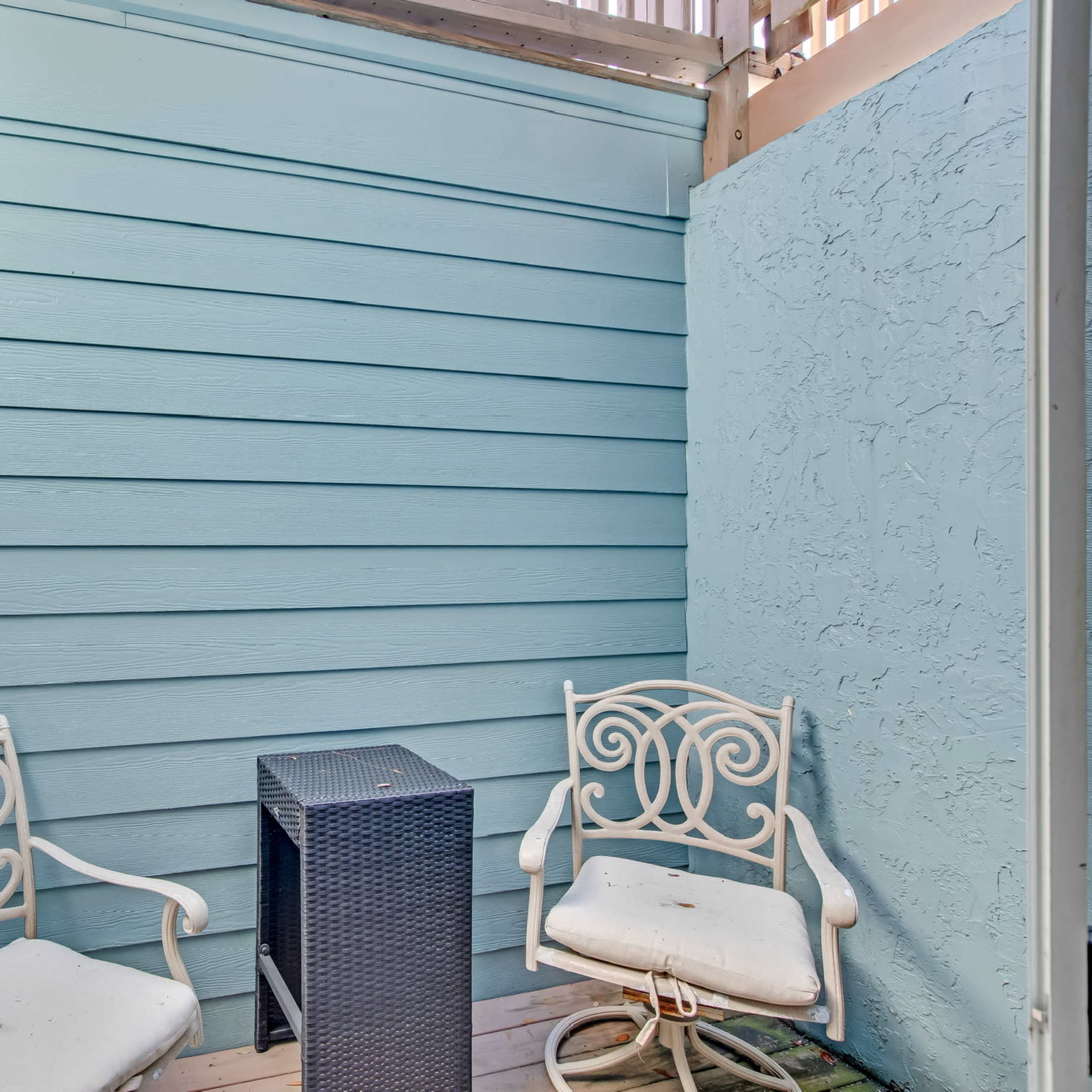 A small patio area with two white metal chairs and a dark wicker side table against blue wooden walls.