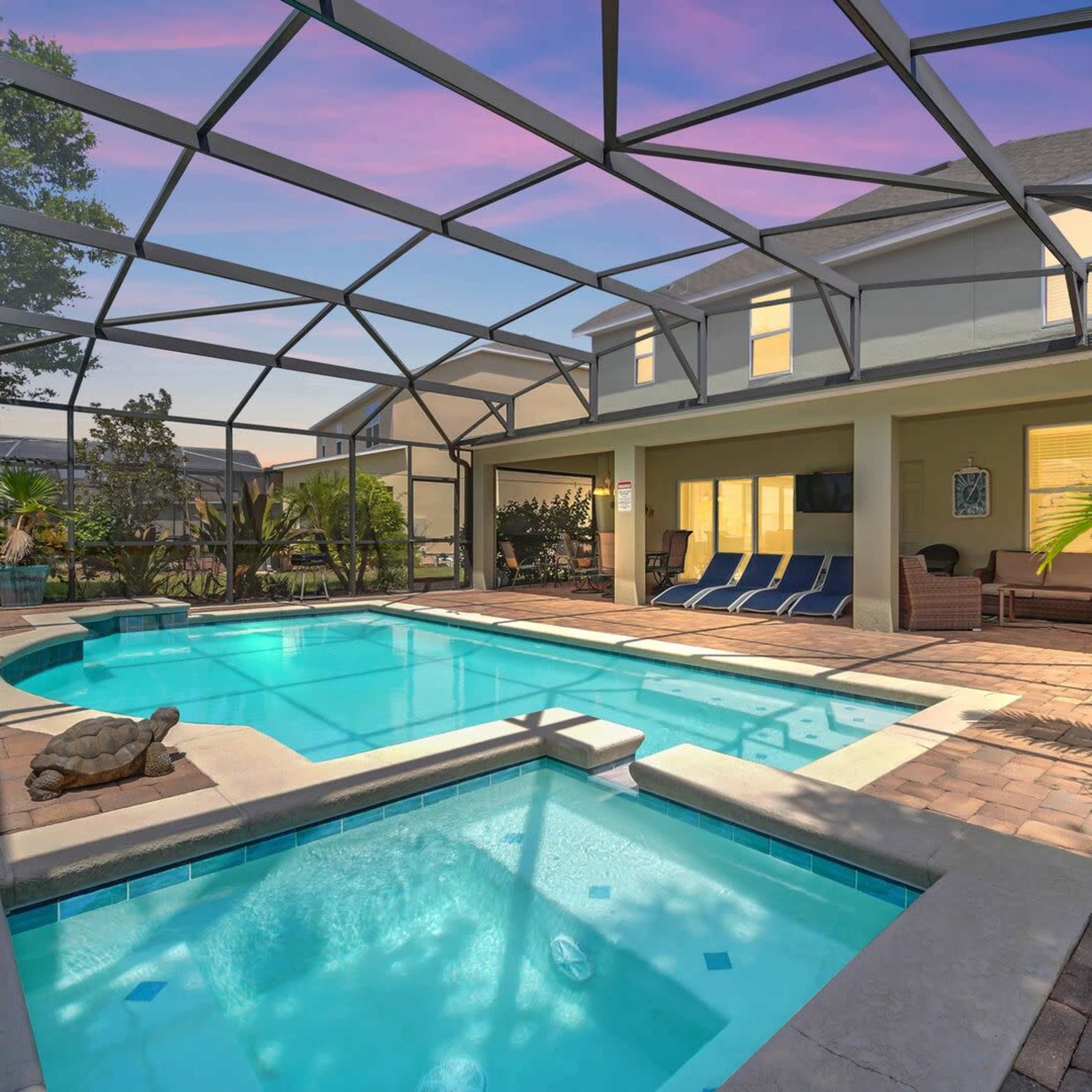 A screened-in pool area with a hot tub, lounge chairs, and tropical plants surrounding the pool.