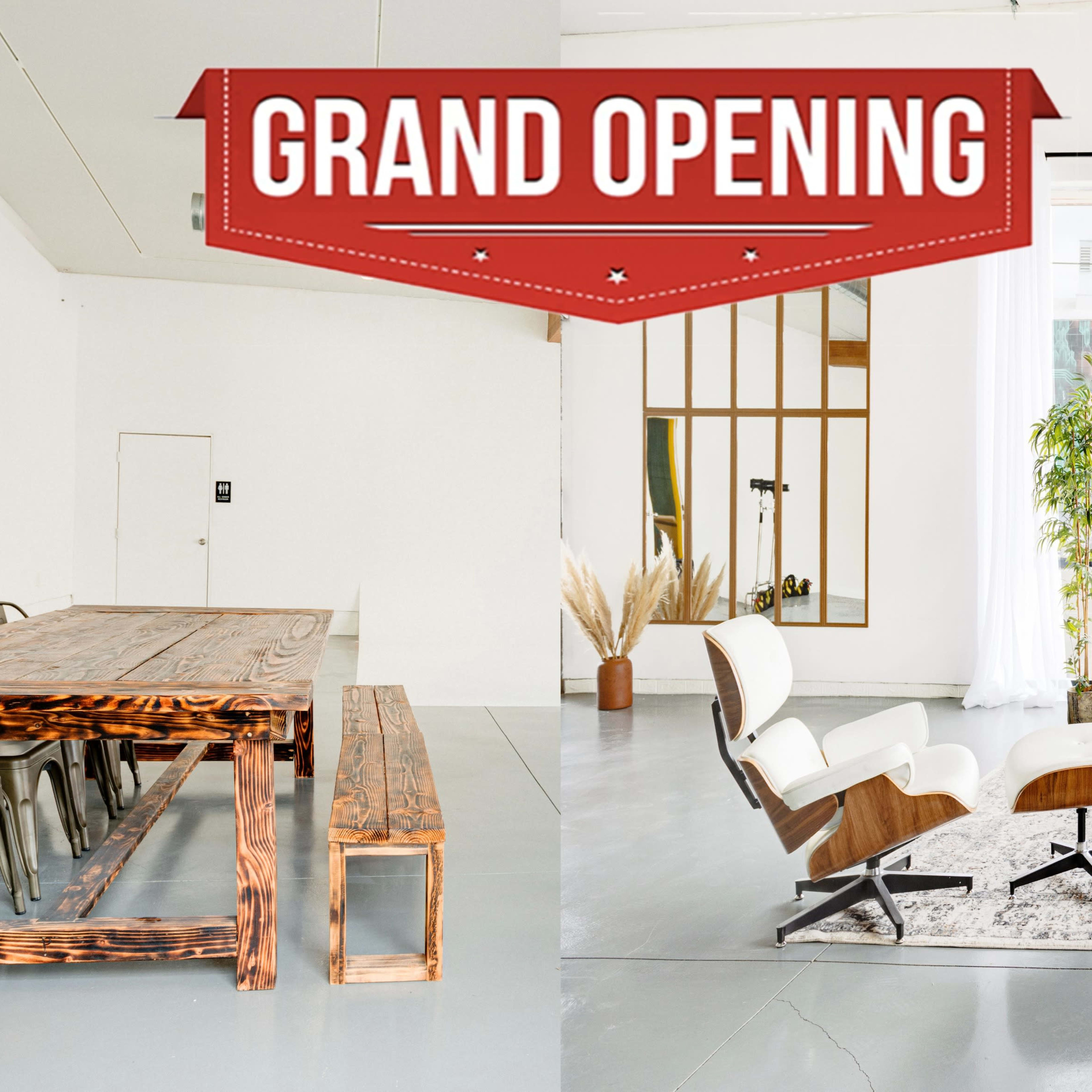 A spacious, modern interior featuring a wooden dining table and benches on one side, and a cozy seating area with a leather couch, coffee table, and large windows on the other, adorned with a "Grand Opening" promotional banner.
