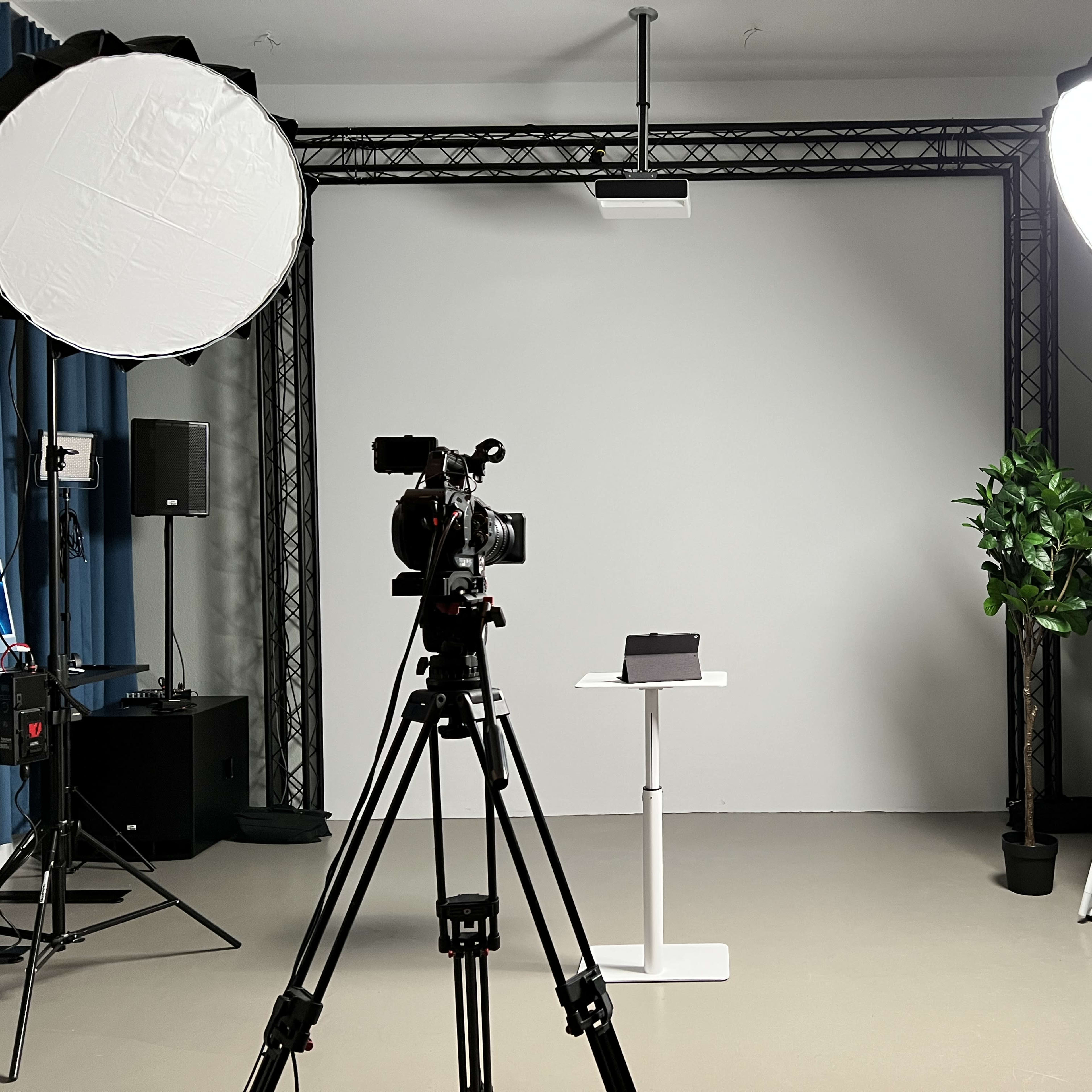 A photography studio setup features two cameras on tripods, a white backdrop, lighting equipment, and a small table with a plant and a black object on it.