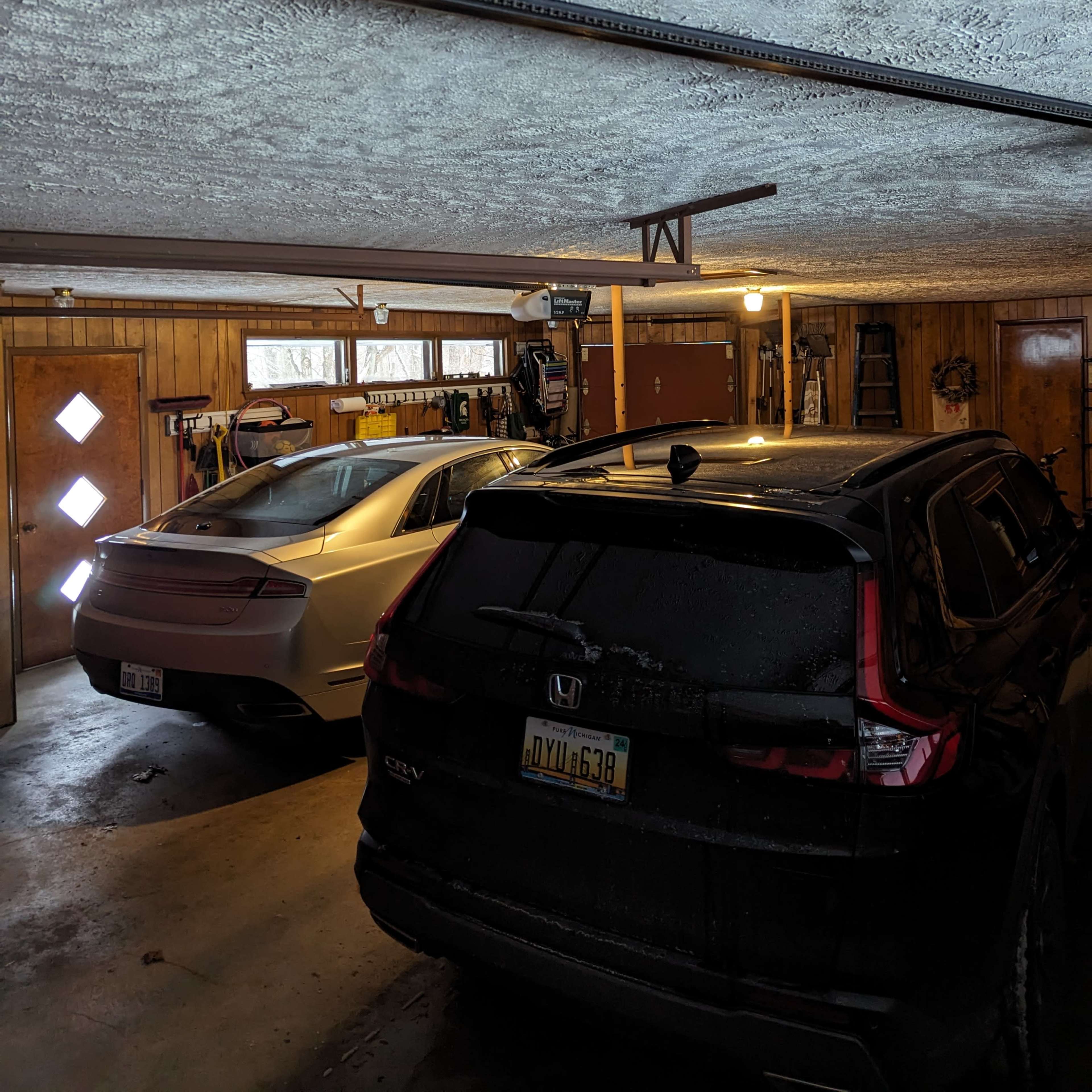 A two-car garage features a white vehicle on the left and a black SUV on the right, with wooden walls and various items stored in the background.
