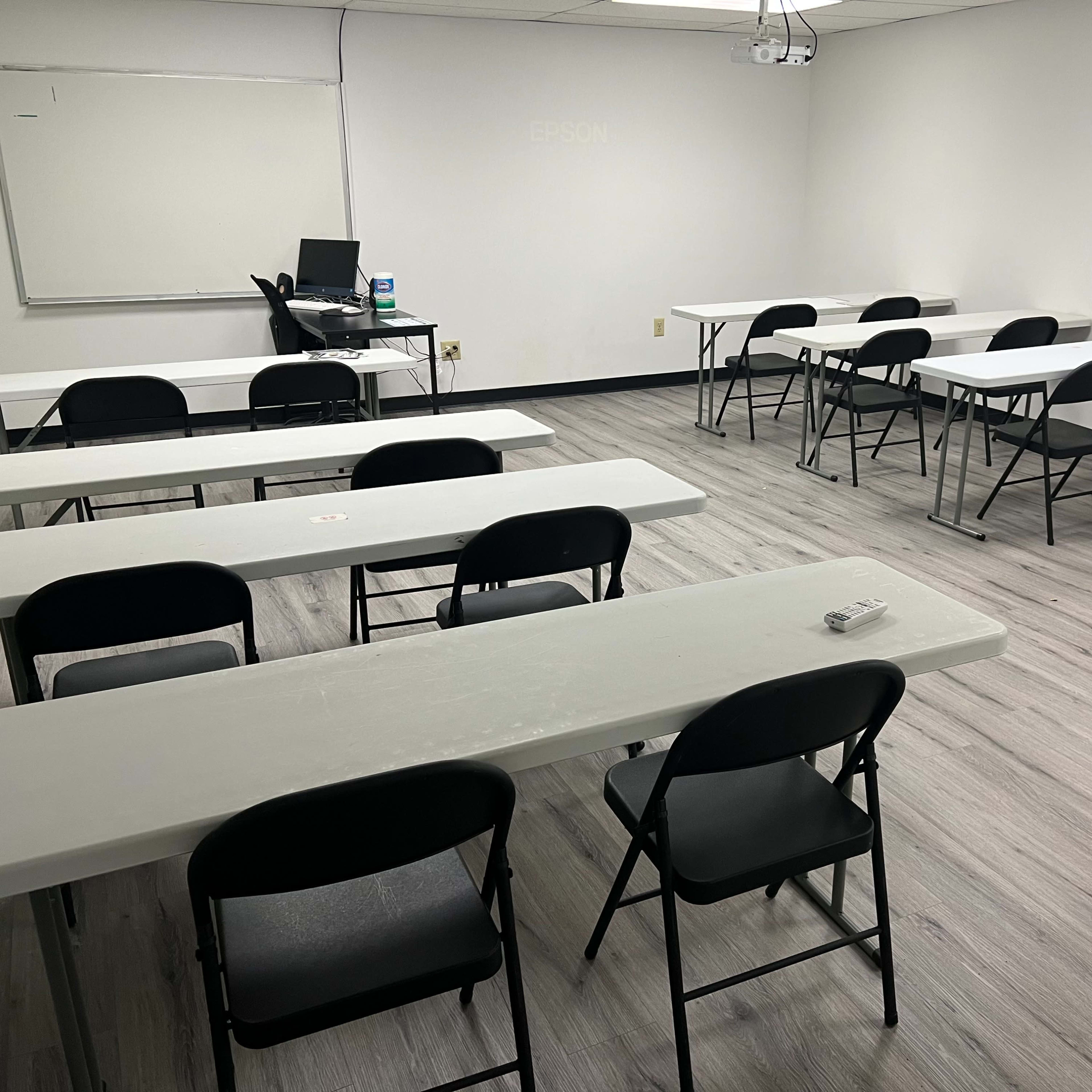 A sparsely furnished classroom with several white tables and black chairs arranged for use.