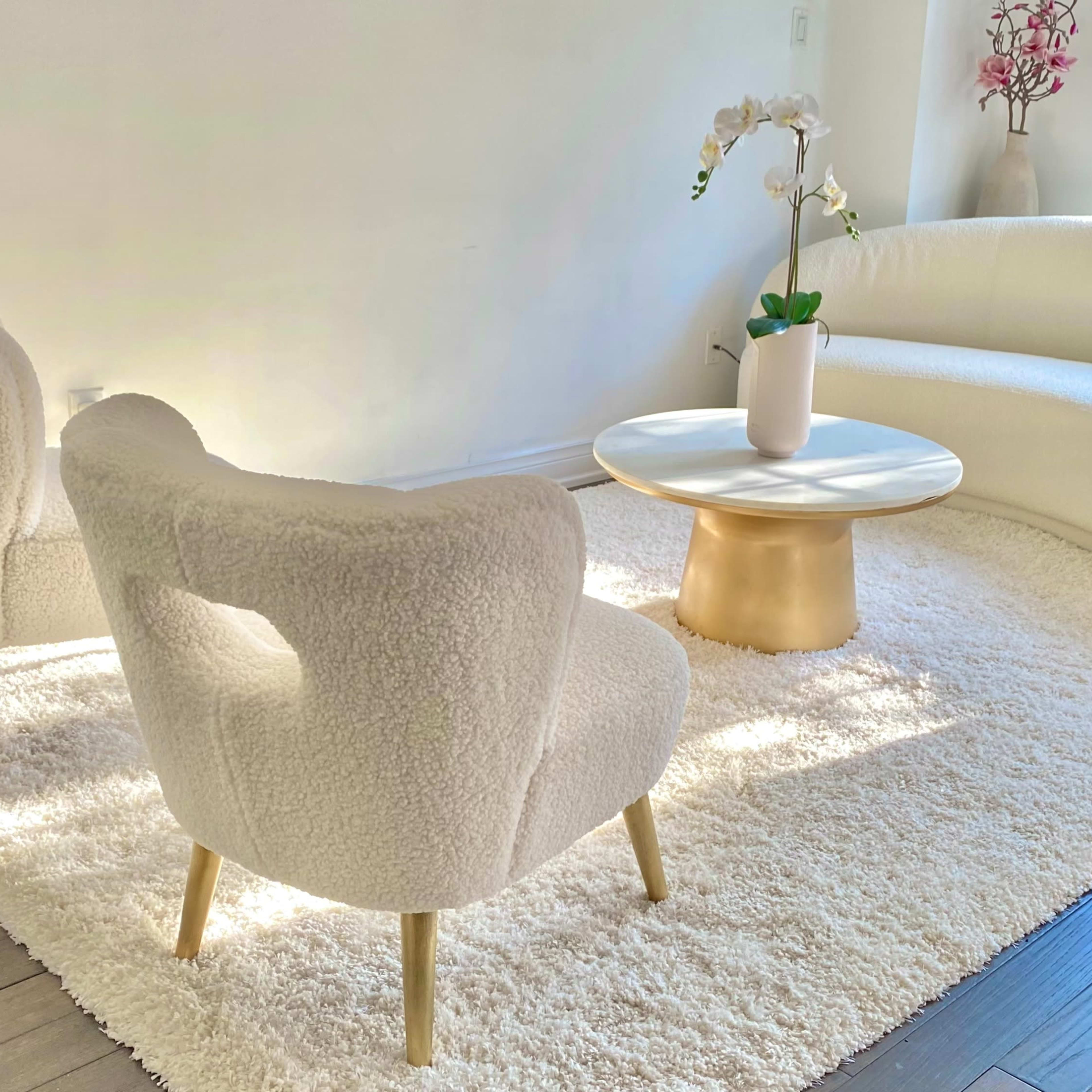 A modern living room features a curved white sofa, two textured chairs, a round table, and an orchid centerpiece on a plush area rug.