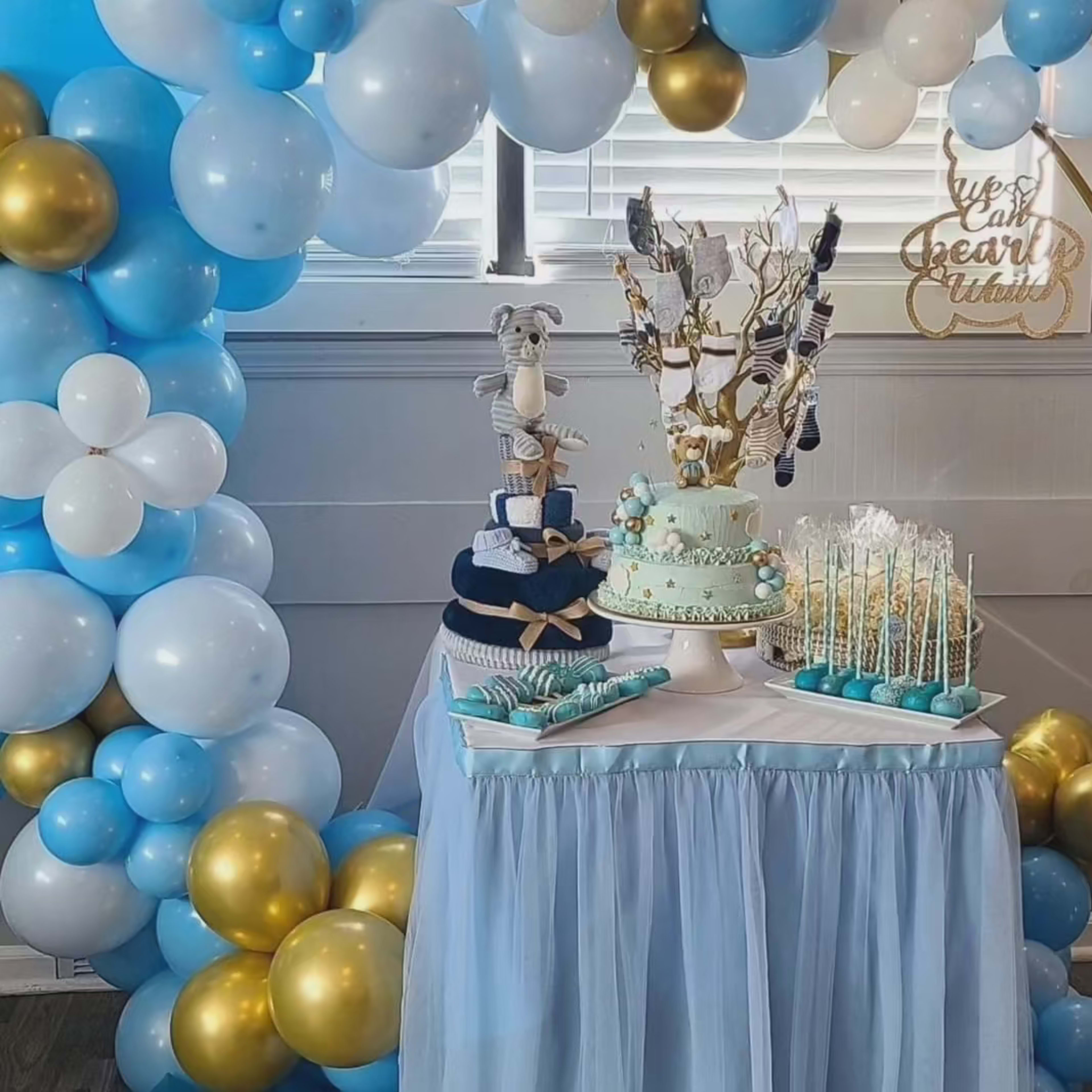 A decorated table displays a multi-tiered cake, blue and gold balloon arrangements, and various sweet treats in a themed setting.