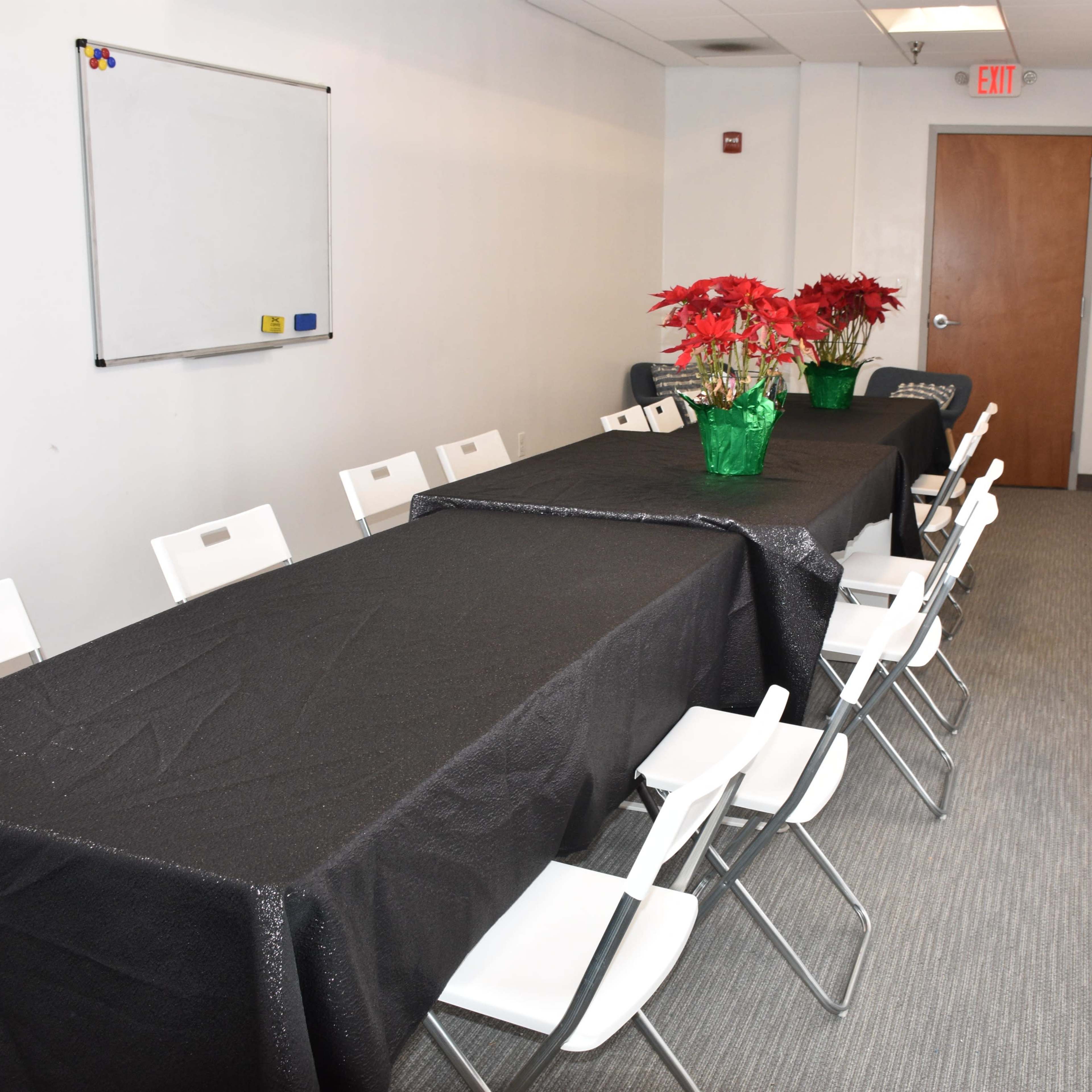 A long table covered with a black tablecloth is set in a conference room, lined with white chairs, and adorned with a centerpiece of potted red poinsettias.