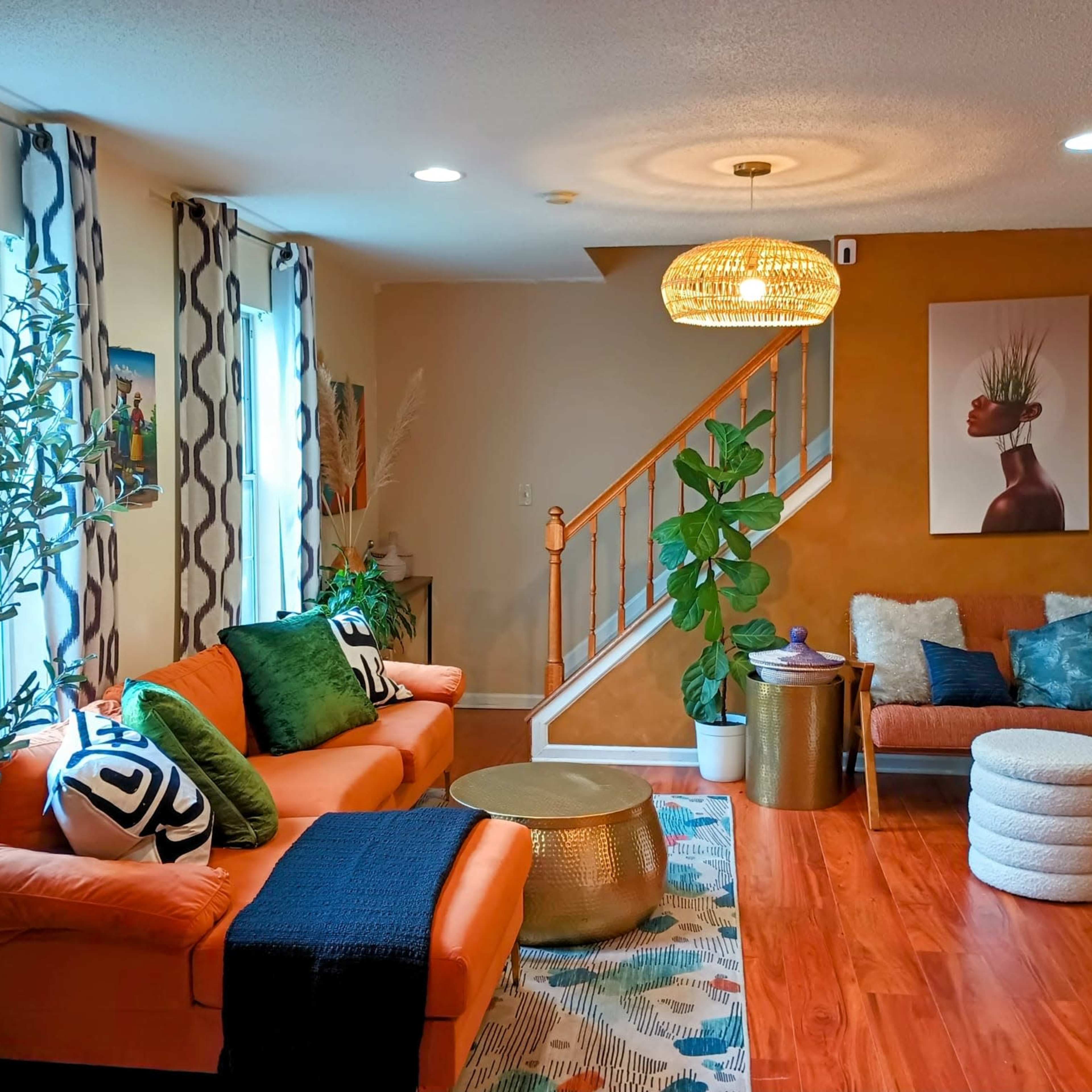 A cozy living room features orange sofas, a round coffee table, and a staircase with a pendant light, surrounded by plants and decorative artwork.