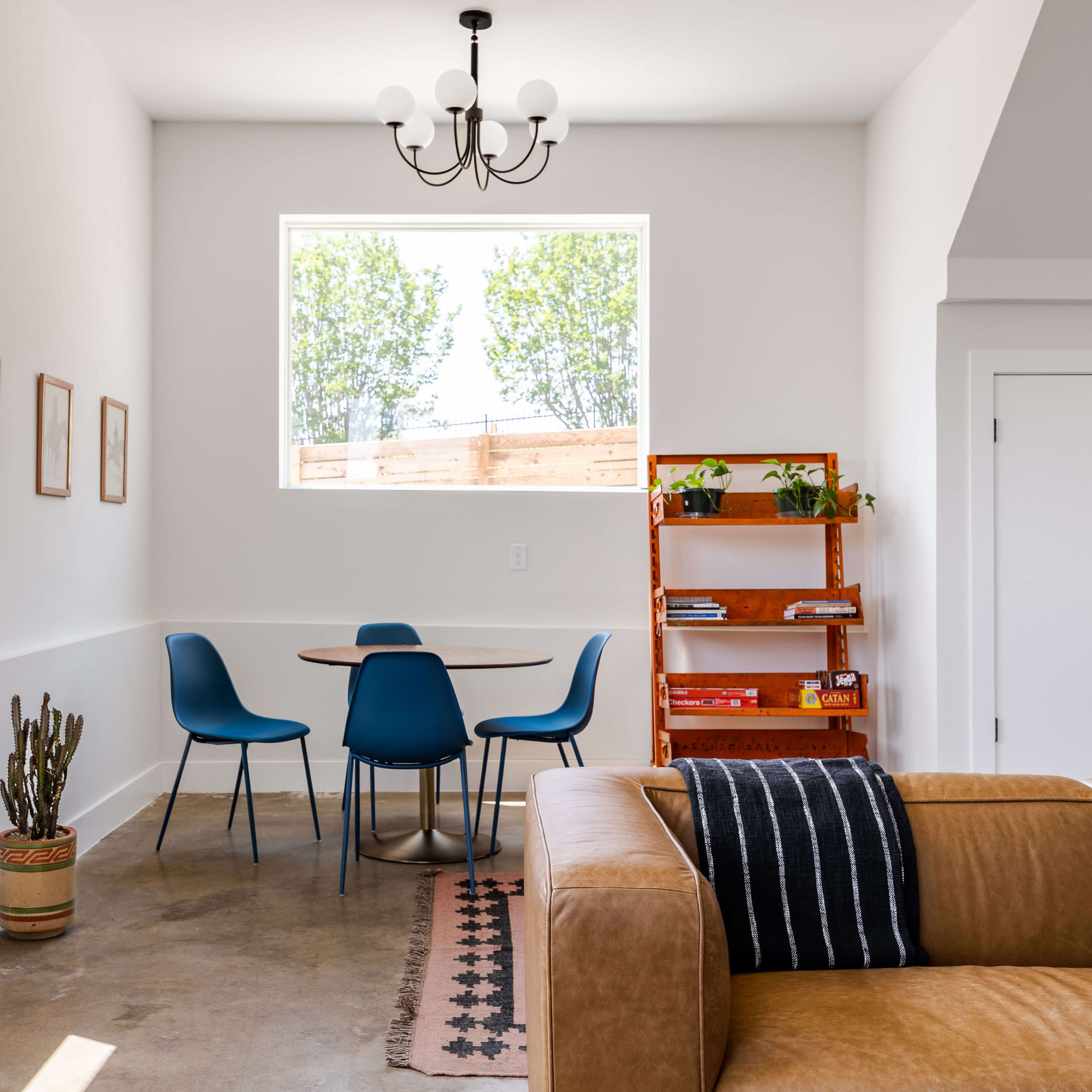 A modern living space featuring a brown leather couch, a round dining table with blue chairs, and a wooden shelving unit with plants and books.