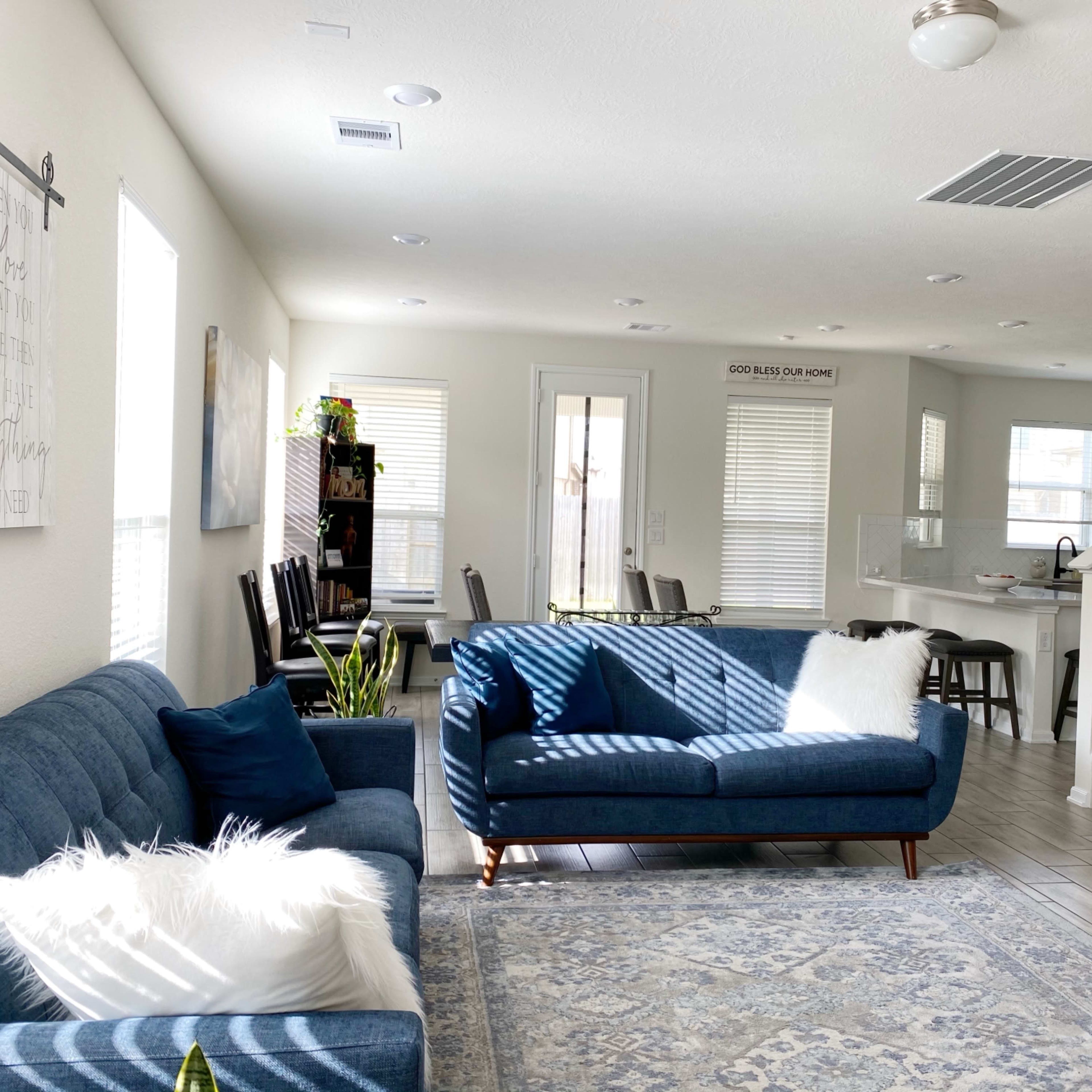 A bright and spacious living room with two blue sofas, a decorative rug, and an open floor plan leading to a dining area and kitchen.