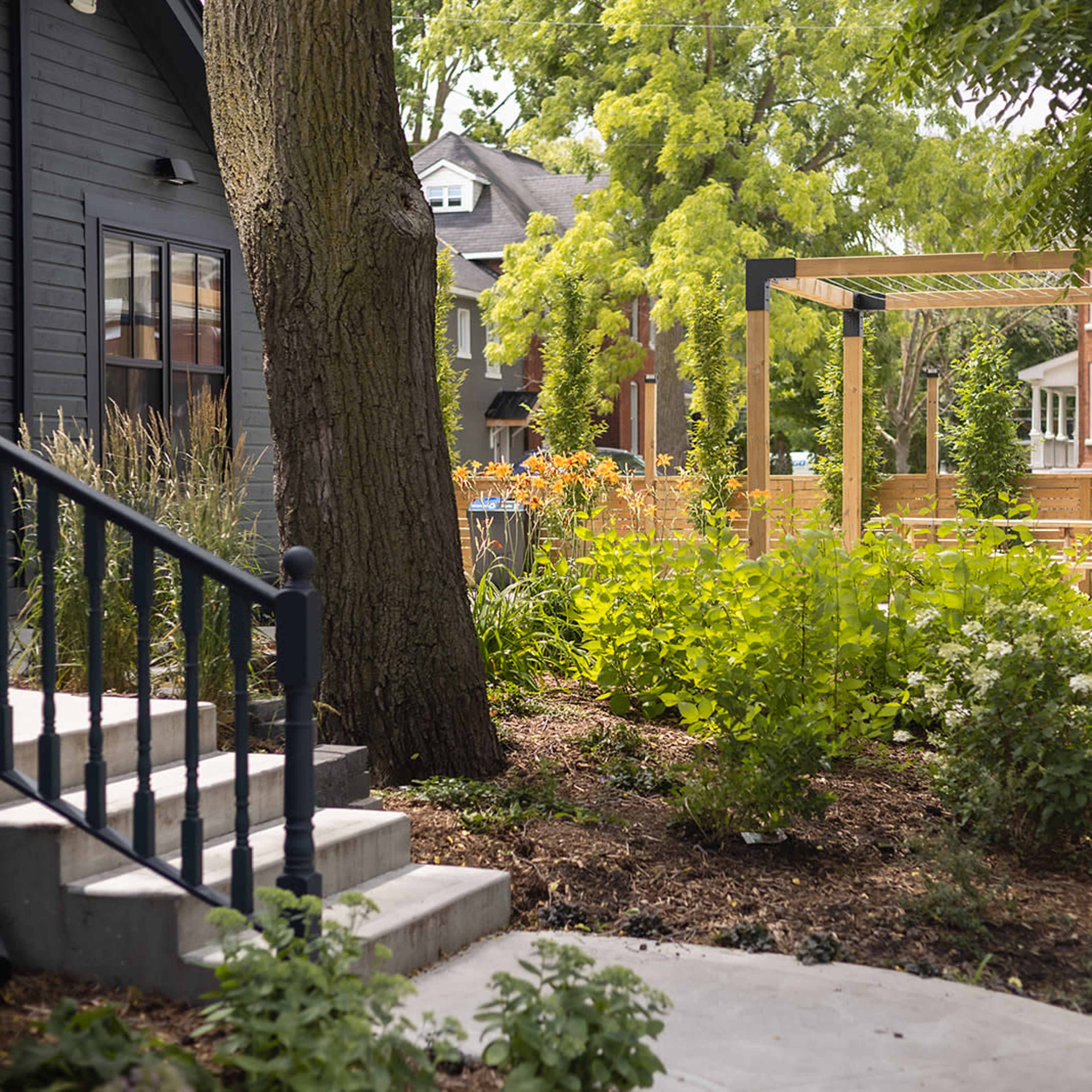 A pathway curves through a landscaped garden featuring greenery and wooden structures beside a dark-colored house.