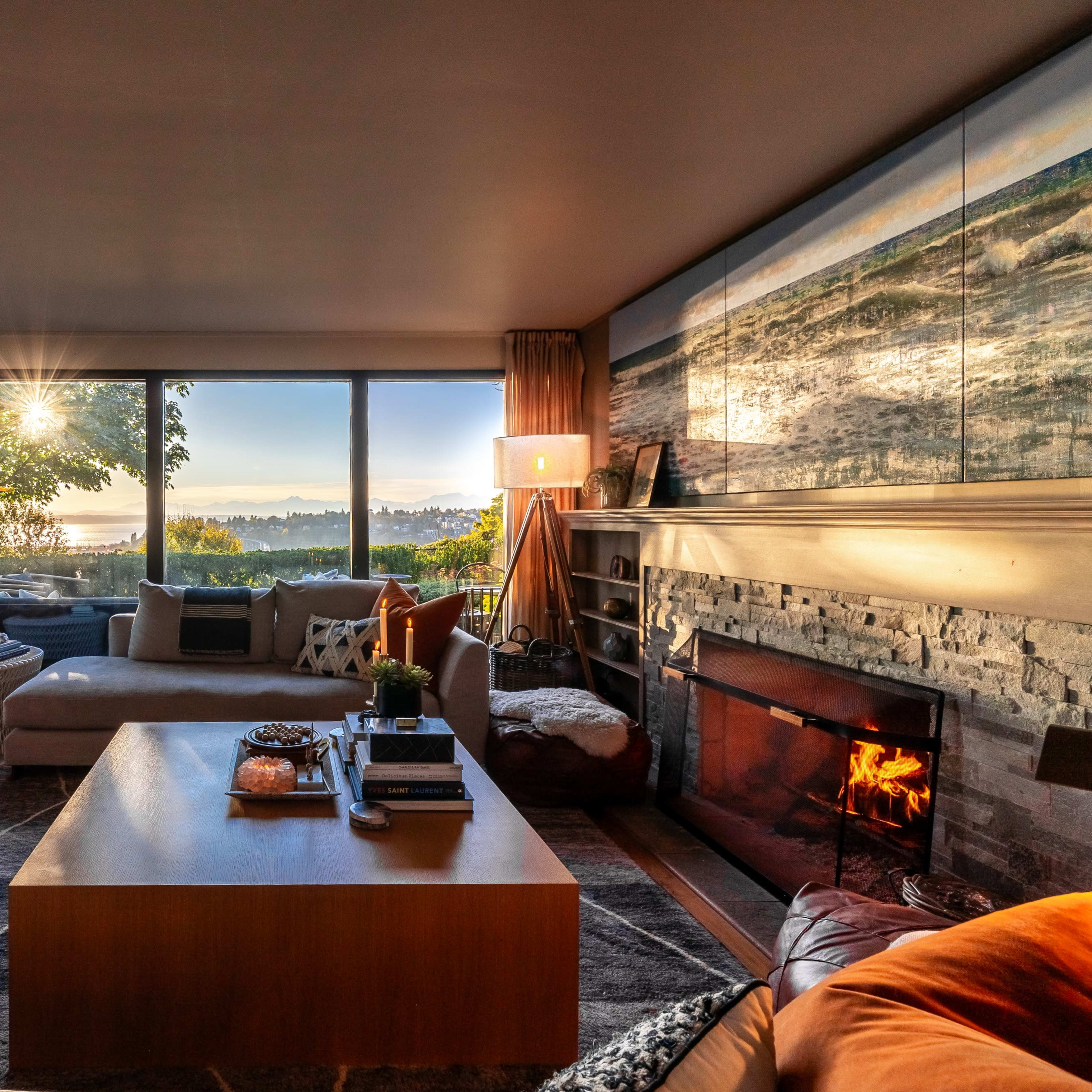A cozy living room features a fireplace, large windows with a view of trees and a sunset, and modern furniture arranged around a coffee table.