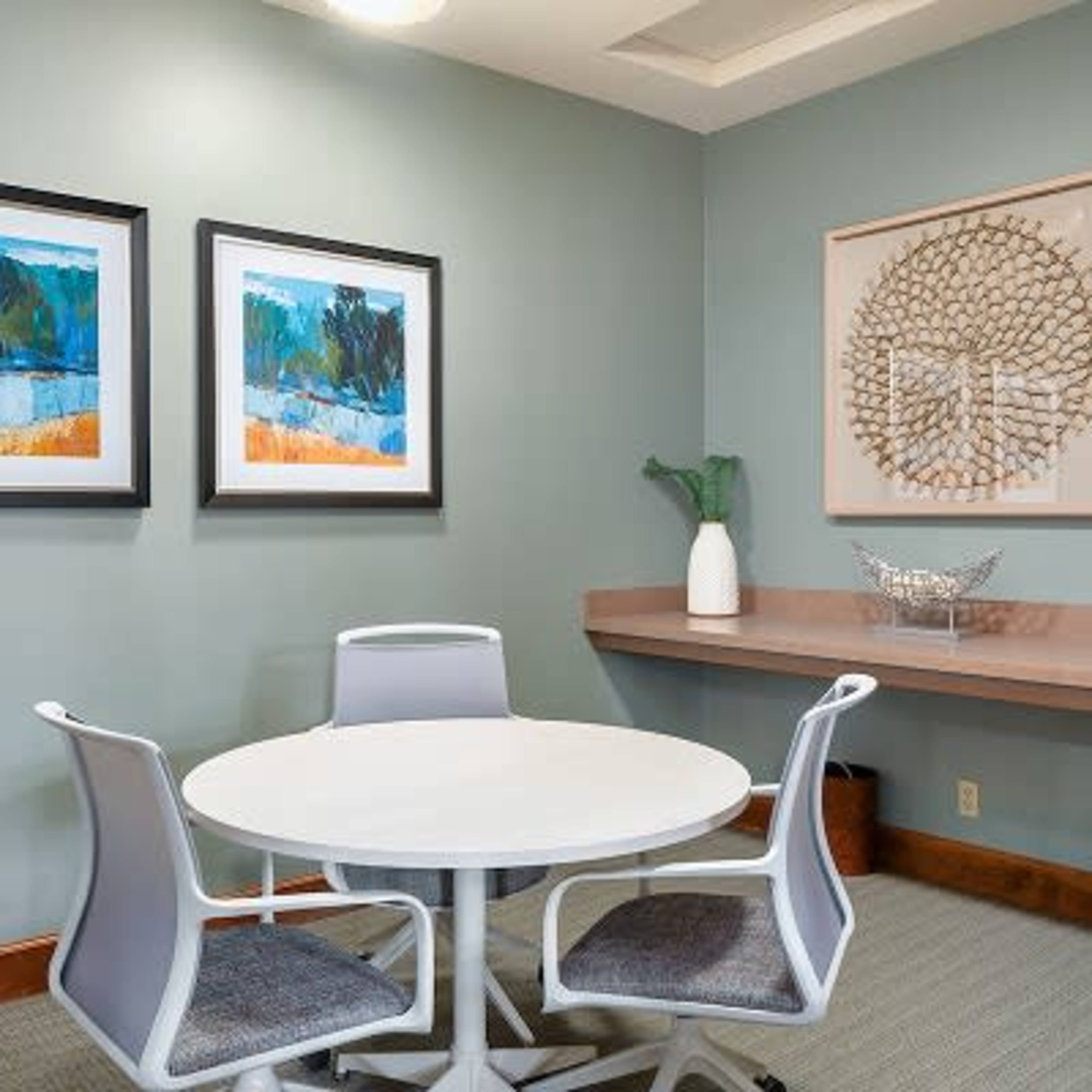 A small meeting room featuring a round white table surrounded by three modern chairs and decorated with framed artworks on the walls.